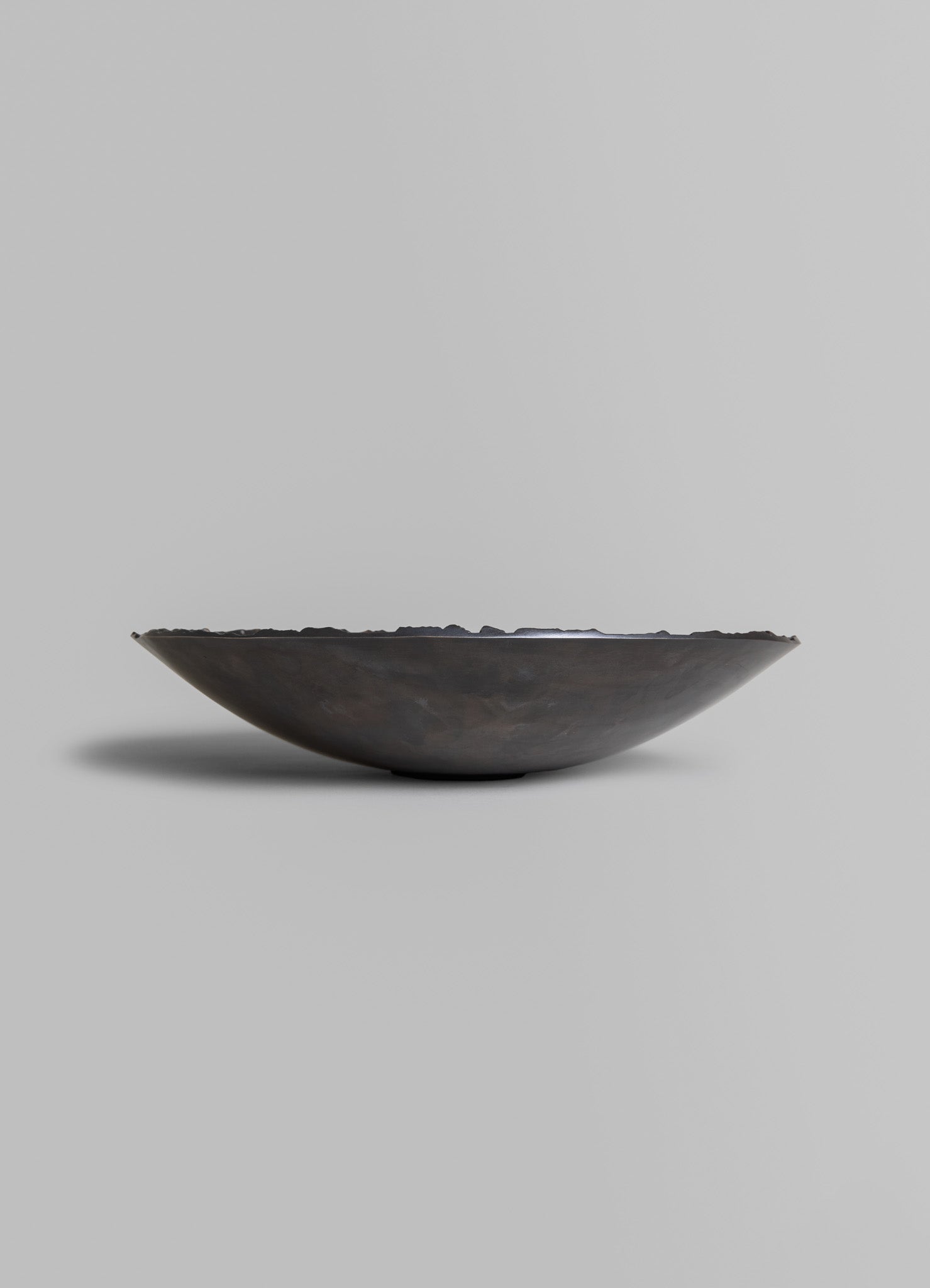 A shallow bowl made of dark, possibly metal or ceramic material with a textured, uneven edge, set against a plain background.