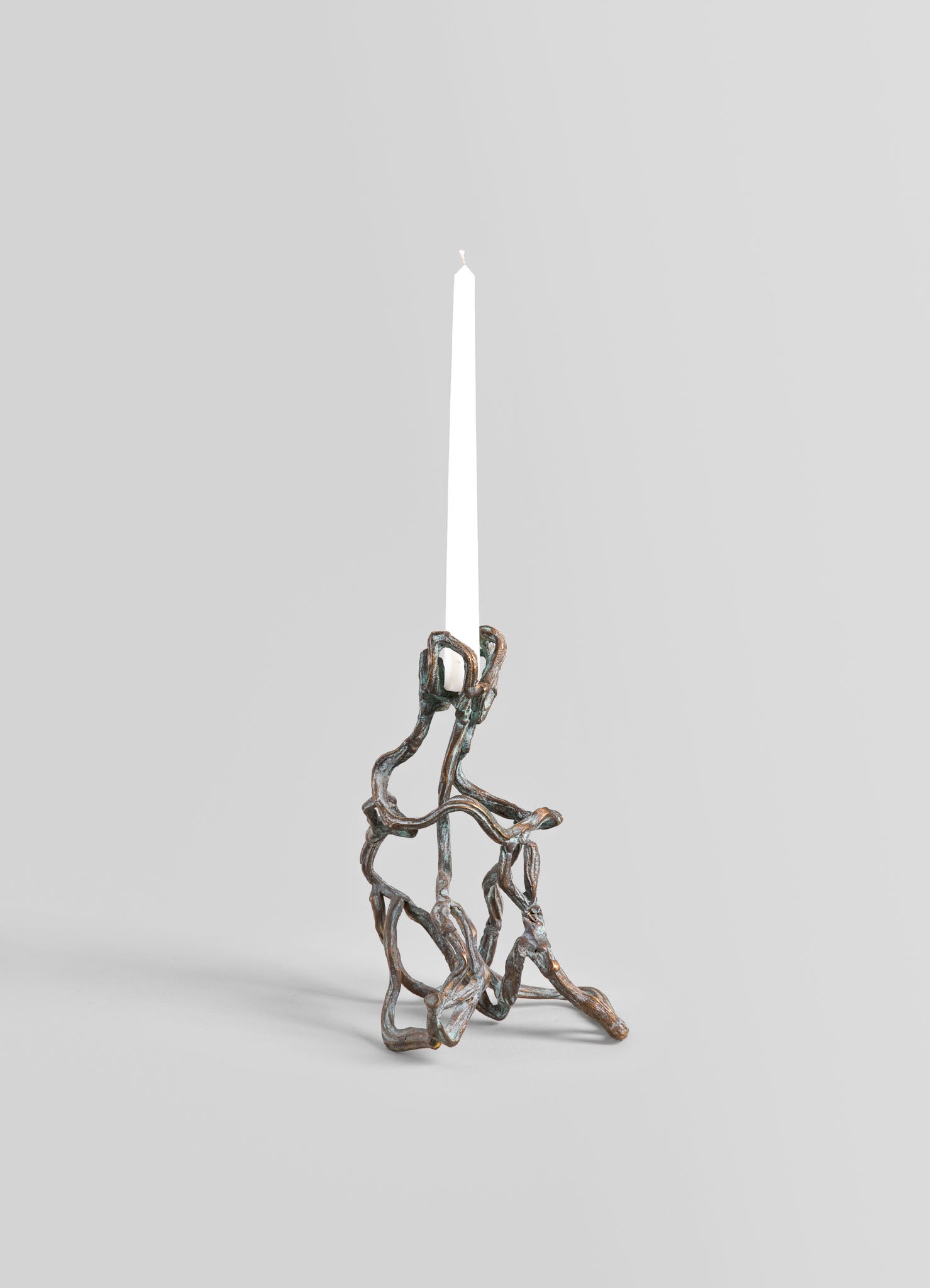 A metal candle holder with an organic, twisted design in a bronze or oxidized finish, holding a tall white taper candle against a neutral background.