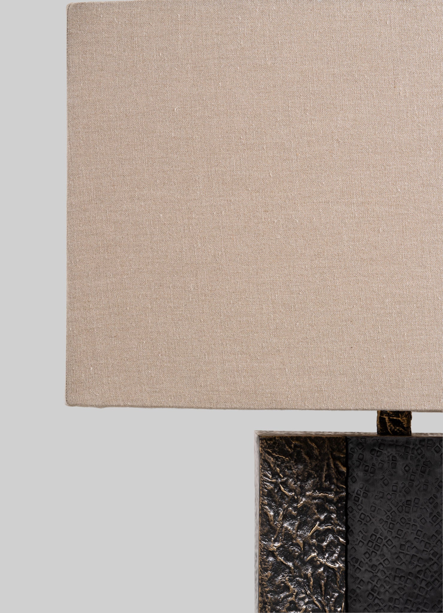 A beige fabric lampshade with a textured surface, mounted on a base with a dark, carved, and textured surface, possibly made of metal or stone. The design combines natural tones with contrasting dark accents, exhibiting a contemporary style.