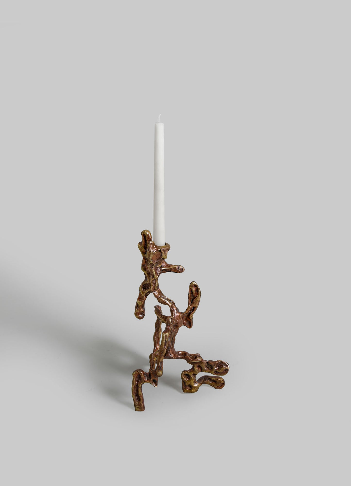 A sculptural candle holder crafted from metal with an organic, irregular shape, featuring a metallic bronze finish, holding a tall white taper candle.