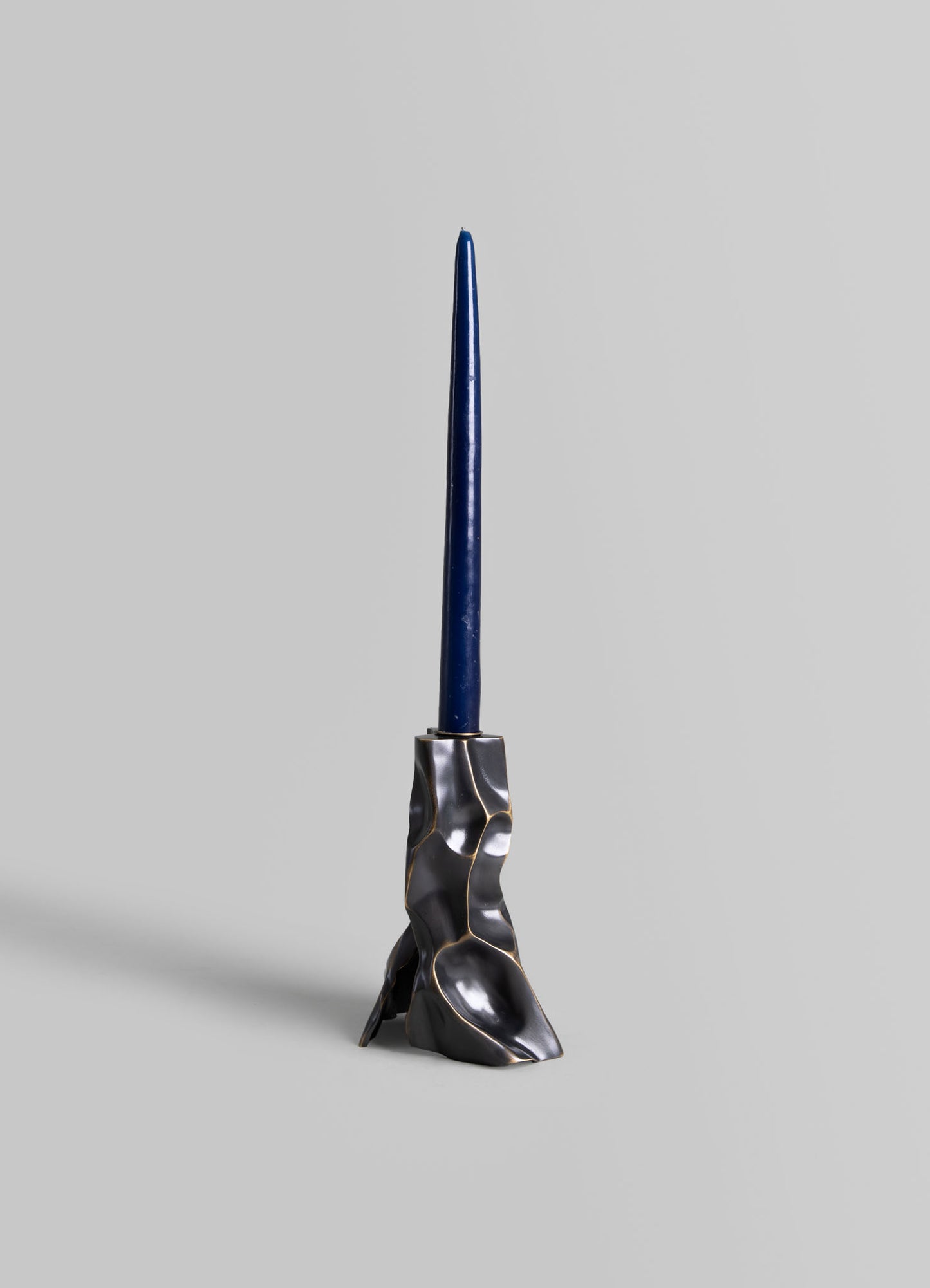 A sculptural candle holder with an irregular, organic-shaped base in metallic silver and black finish, holding a tall, slender blue candle. The design combines modern and handcrafted elements with a contemporary style.