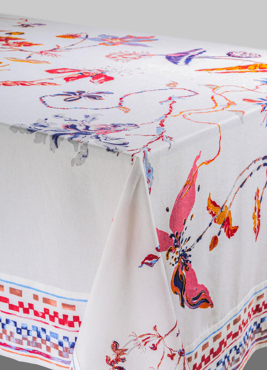 A tablecloth made of white cotton with a colorful floral pattern, featuring pink, purple, orange, and blue flowers and foliage. The border displays a geometric motif in shades of red, blue, and orange.