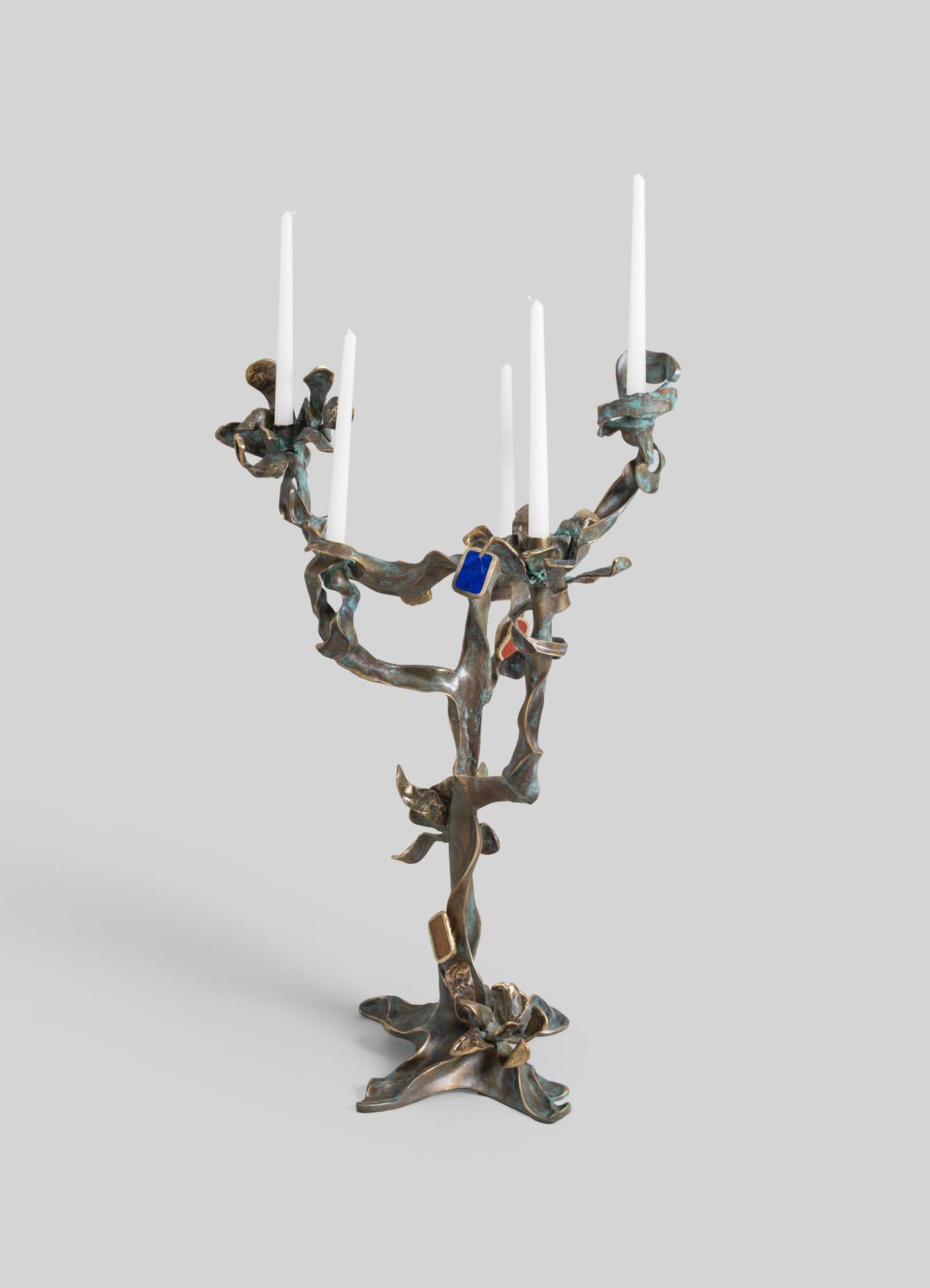 Metallic bronze candle holder with an organic, twisted design resembling a tree branch, featuring five white taper candles and small colored accents. The piece has a contemporary, sculptural style with a rustic, artisanal aesthetic.