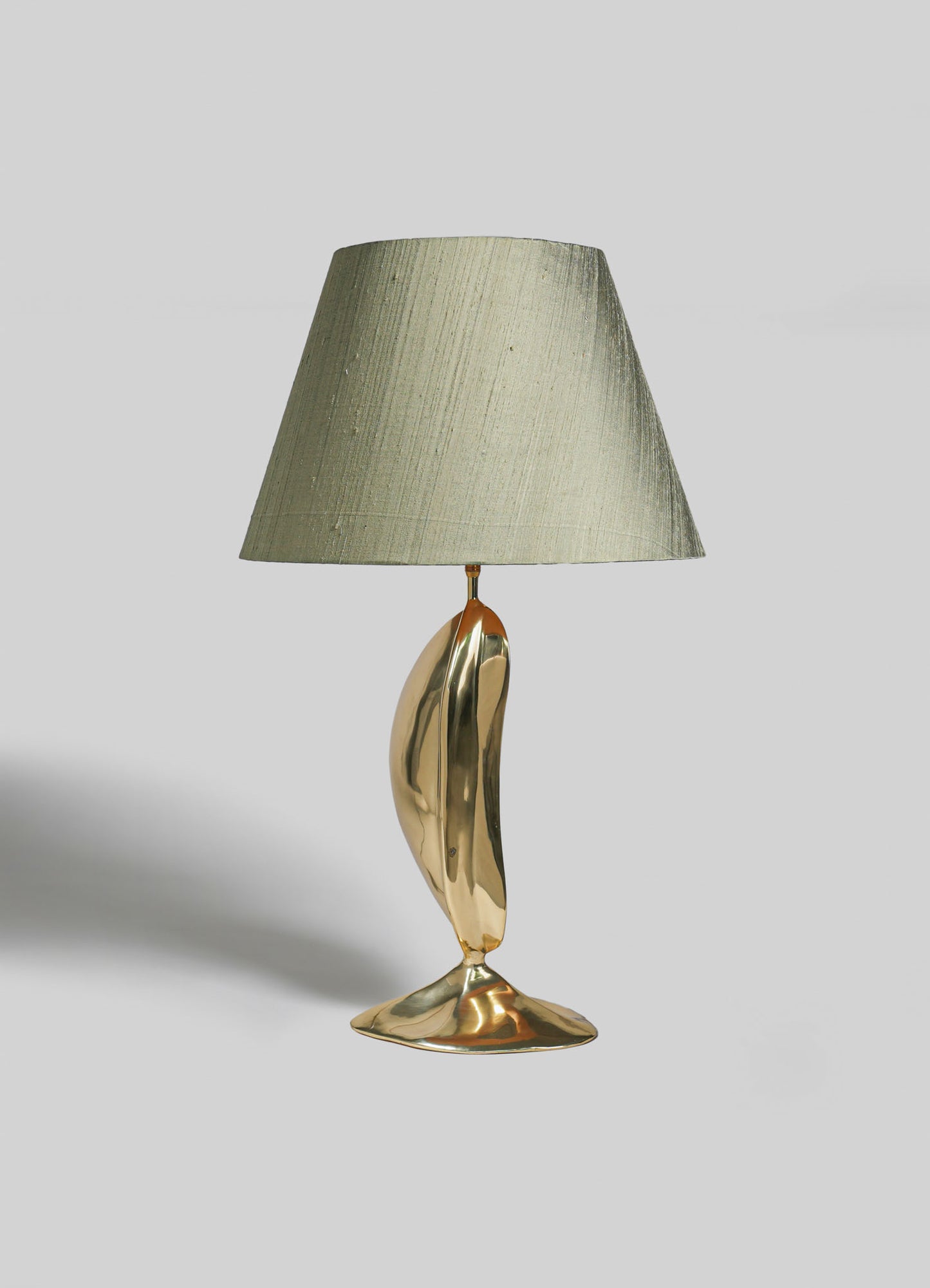 A table lamp with a beige fabric shade and a sculptural metallic gold base, featuring a smooth, reflective surface and an organic, curvilinear design.