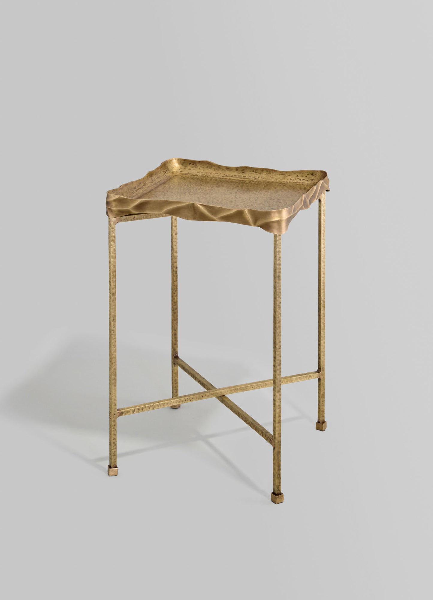 A brass side table with a hammered finish and a square, textured tray top with a slightly ruffled edge, supported by slender, tapered legs with crossbars for stability.
