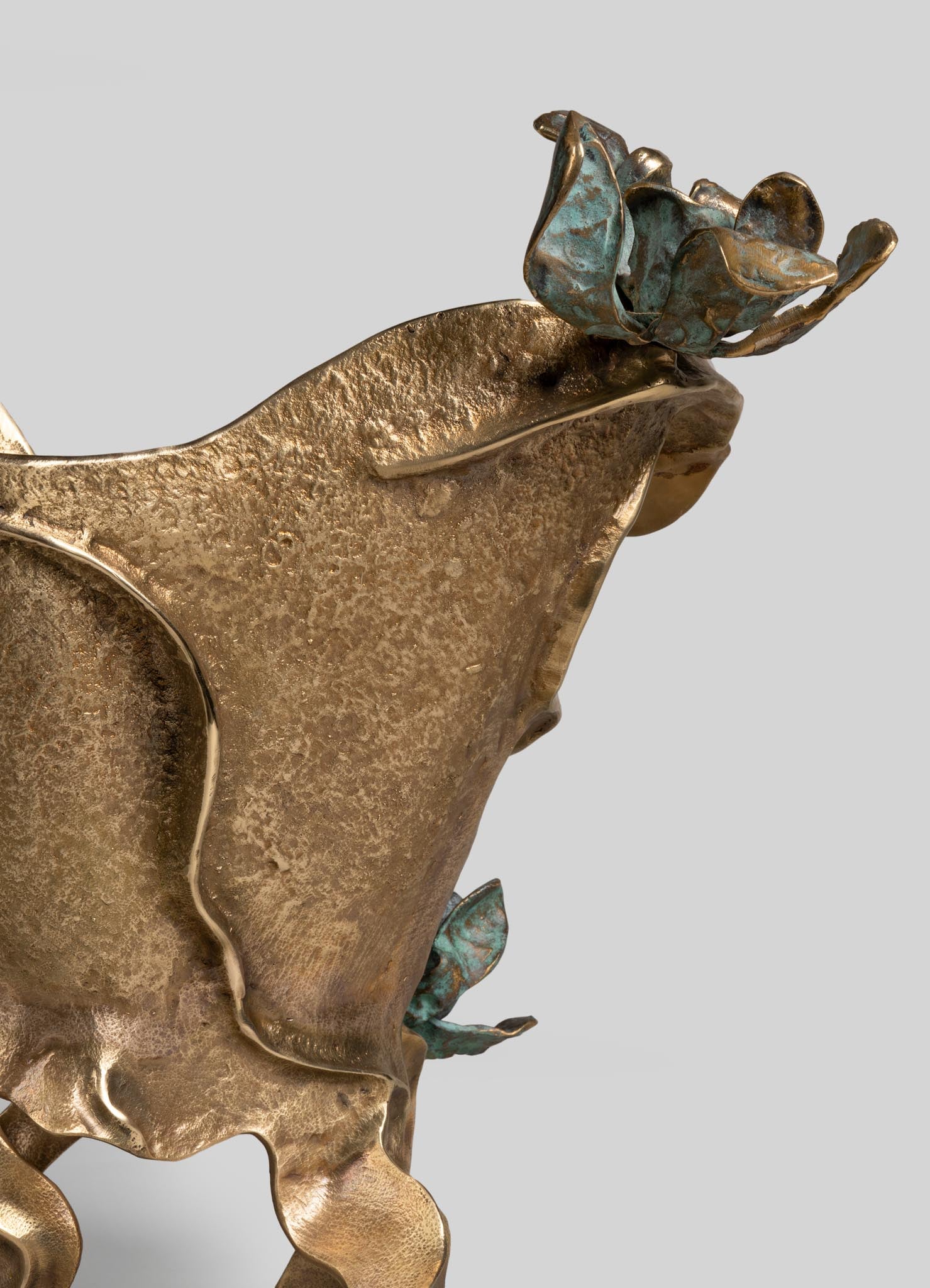 A decorative metal sculpture featuring a textured brass or bronze finish with floral motifs, including detailed petal and leaf shapes. The piece has an artistic, handcrafted style with a combination of warm metallic tones and hints of patina.