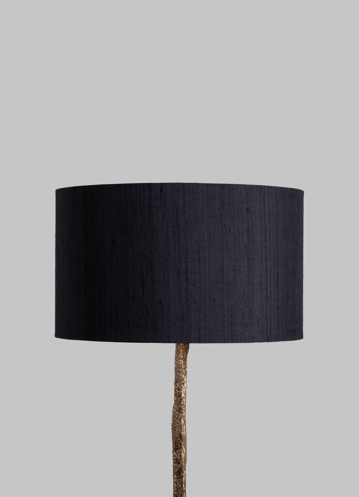 A floor lamp with a cylindrical black fabric lampshade and a textured gold-colored metal stand, set against a plain light gray background.