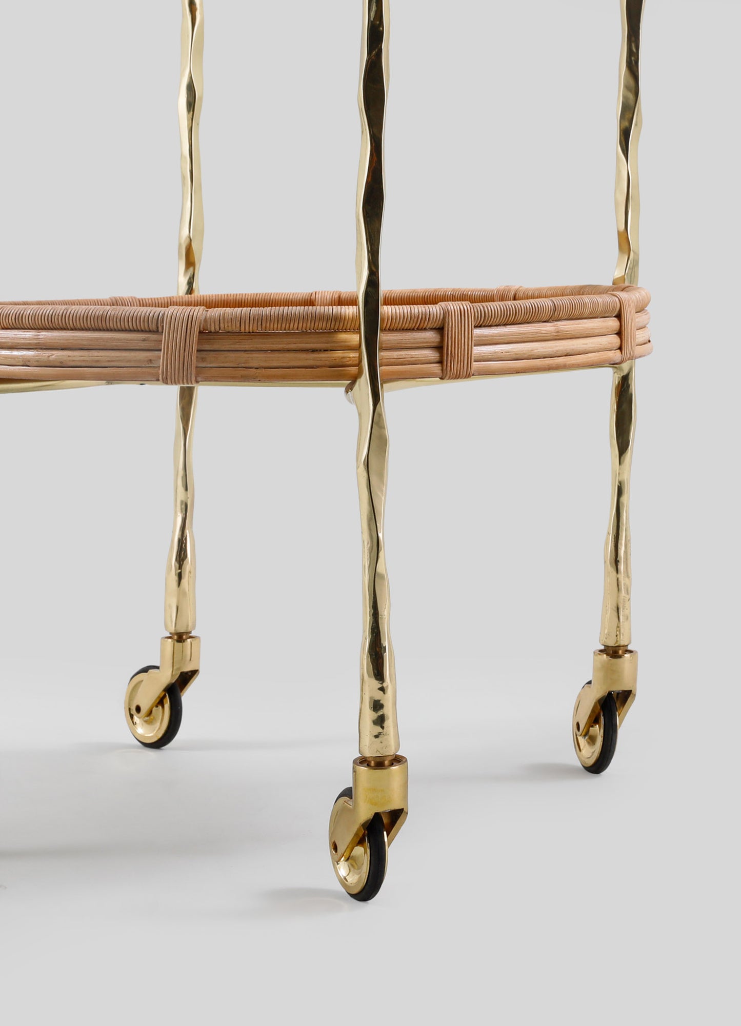 Close-up of the lower part of VIYA's Assam Brass & Cane Trolley, showing four gold-colored forged brass legs with small wheels and a round woven cane shelf near the base.