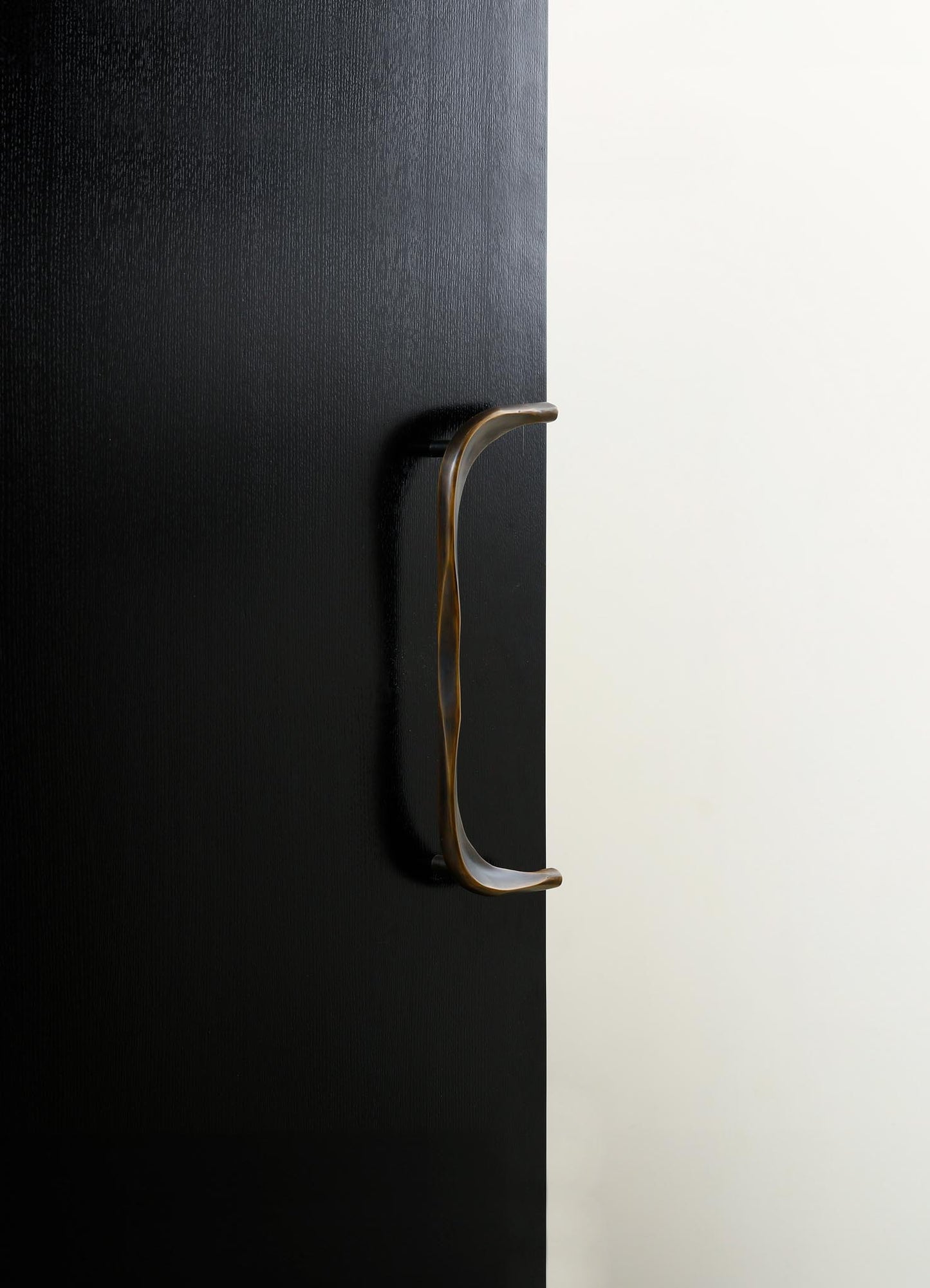 A close-up of a black textured door with a curved metal handle in a bronze or brass finish. The door has a minimalist design and is set against a plain, light-colored wall.