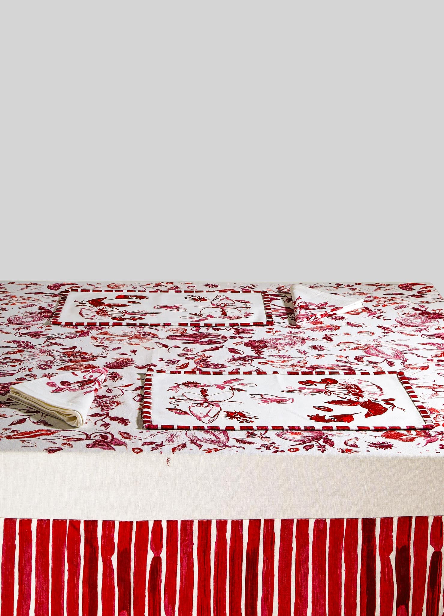 Red and white printed tablecloth with floral and striped motifs, paired with matching napkins and placemats featuring red borders. The fabric appears to be linen or cotton in a contemporary style.