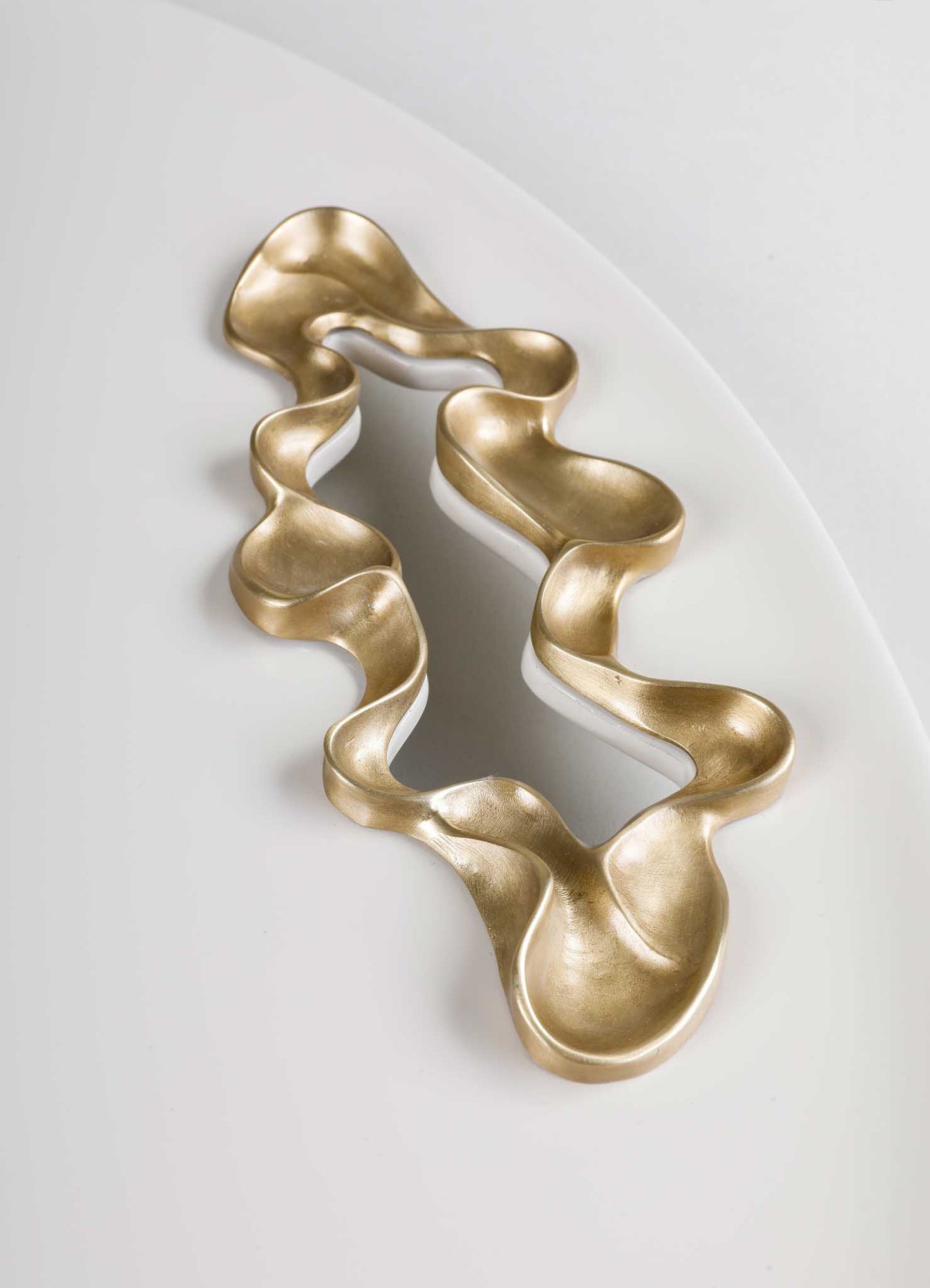 A decorative candle holder made of metal with a flowing, organic shape and a matte gold finish, placed on a white surface. The design features smooth curves resembling a sculptural art piece with no visible handles or additional motifs.