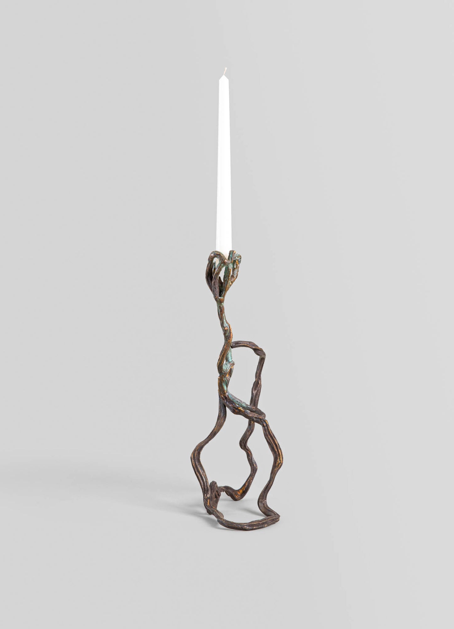 A metallic candle holder with an organic, branch-like design, holding a tall white candle. The finish appears to be bronze or aged metal, set against a plain, light grey background.
