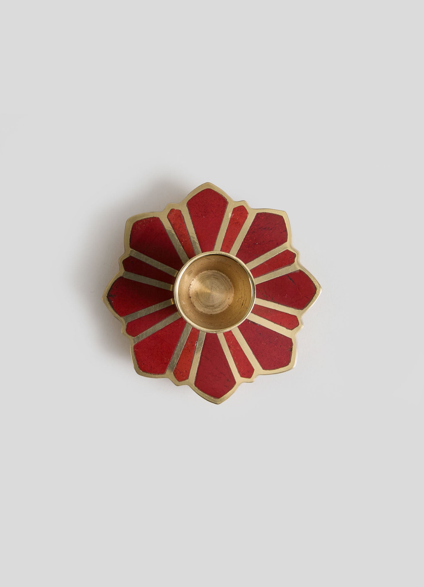 A decorative brass and red lacquered ceramic candle holder with a flower petal design, featuring a circular central bowl for candles, set on a plain surface.