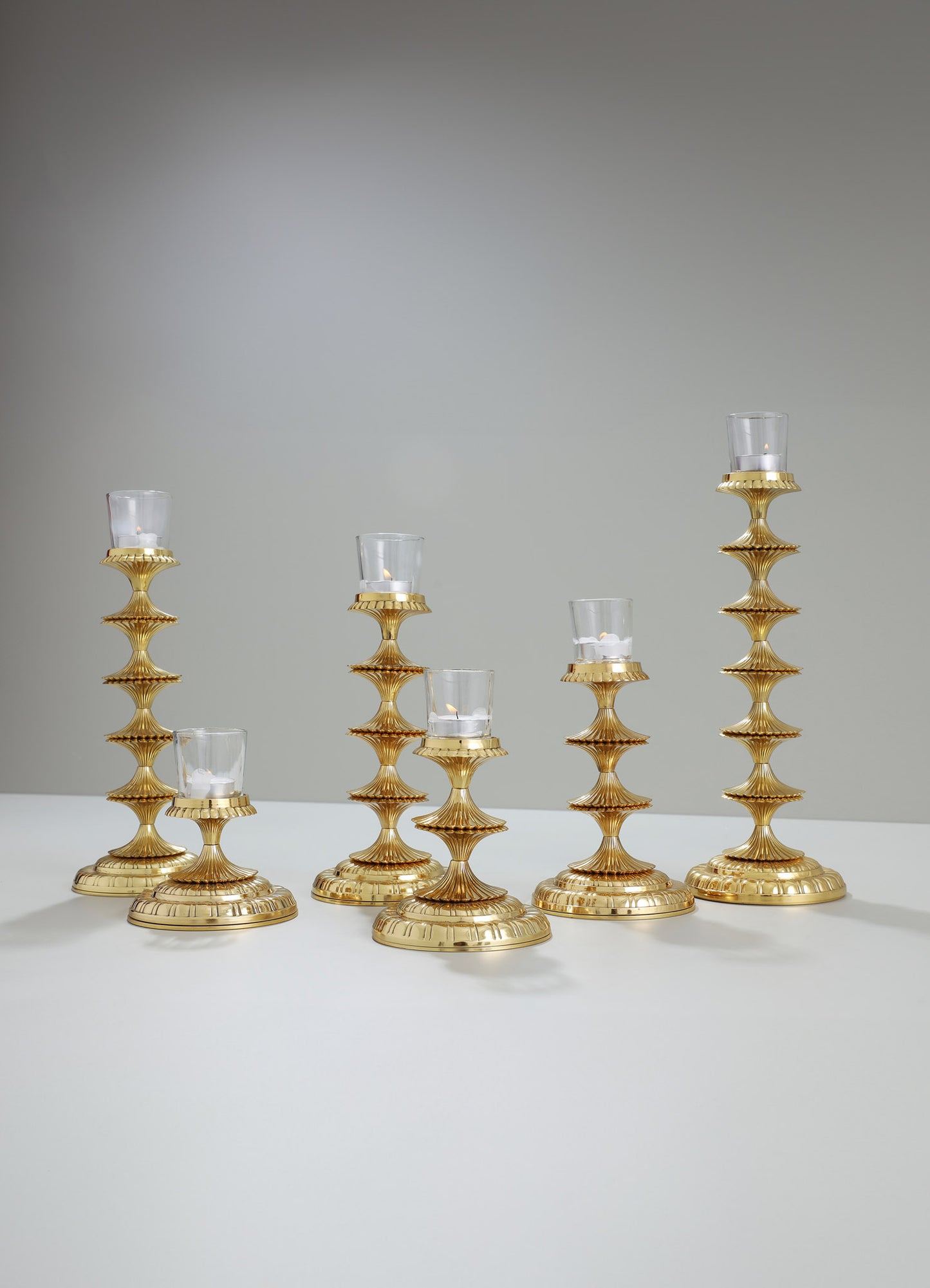 Set of six brass candlesticks with stacked, fluted tiers and a shiny gold finish, each holding a glass holder with a small white candle. The design has a luxurious and contemporary style with elegant, ribbed motifs.