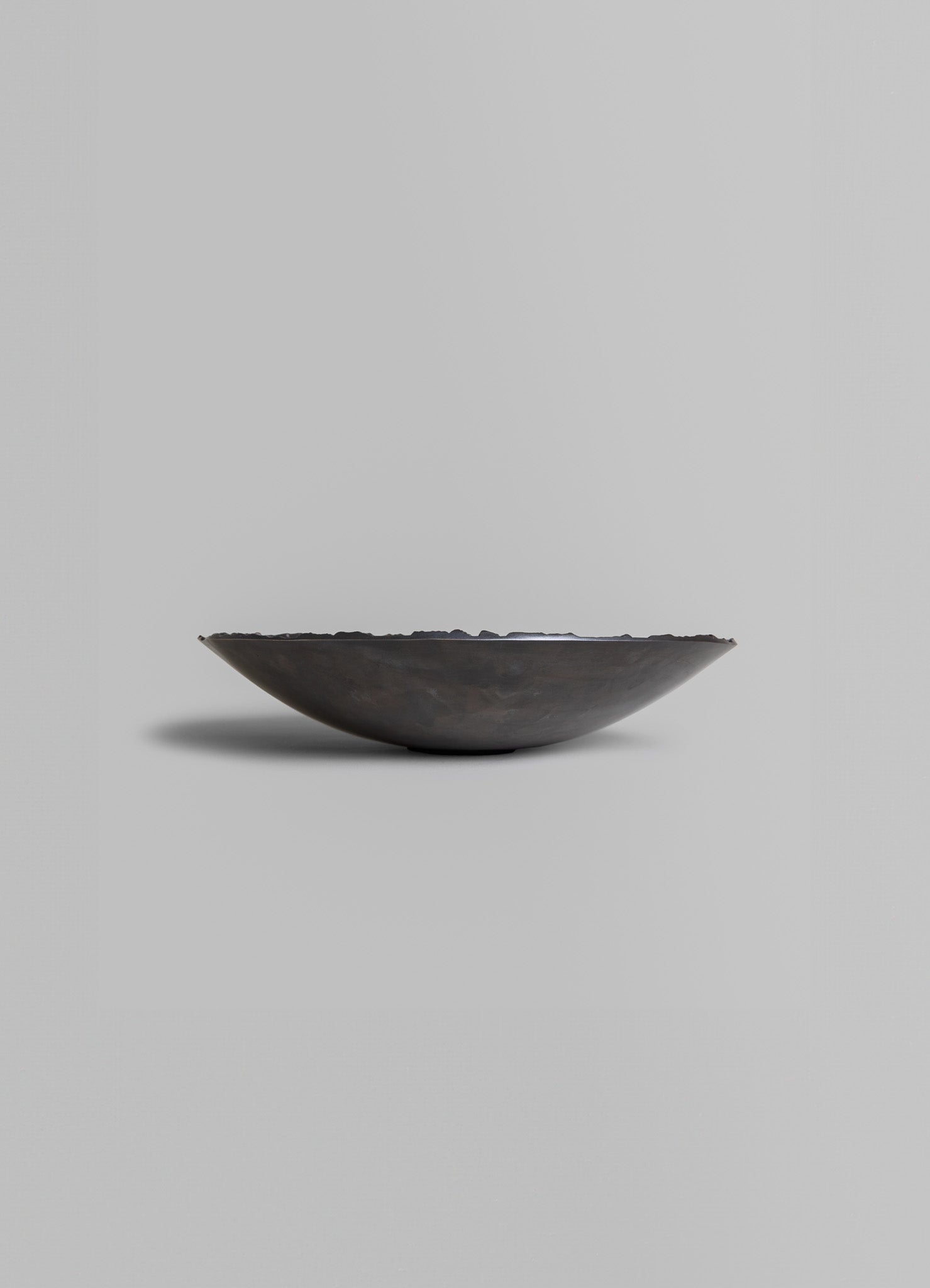 A shallow bowl made of dark metal with an uneven, textured edge, set against a plain light gray background.