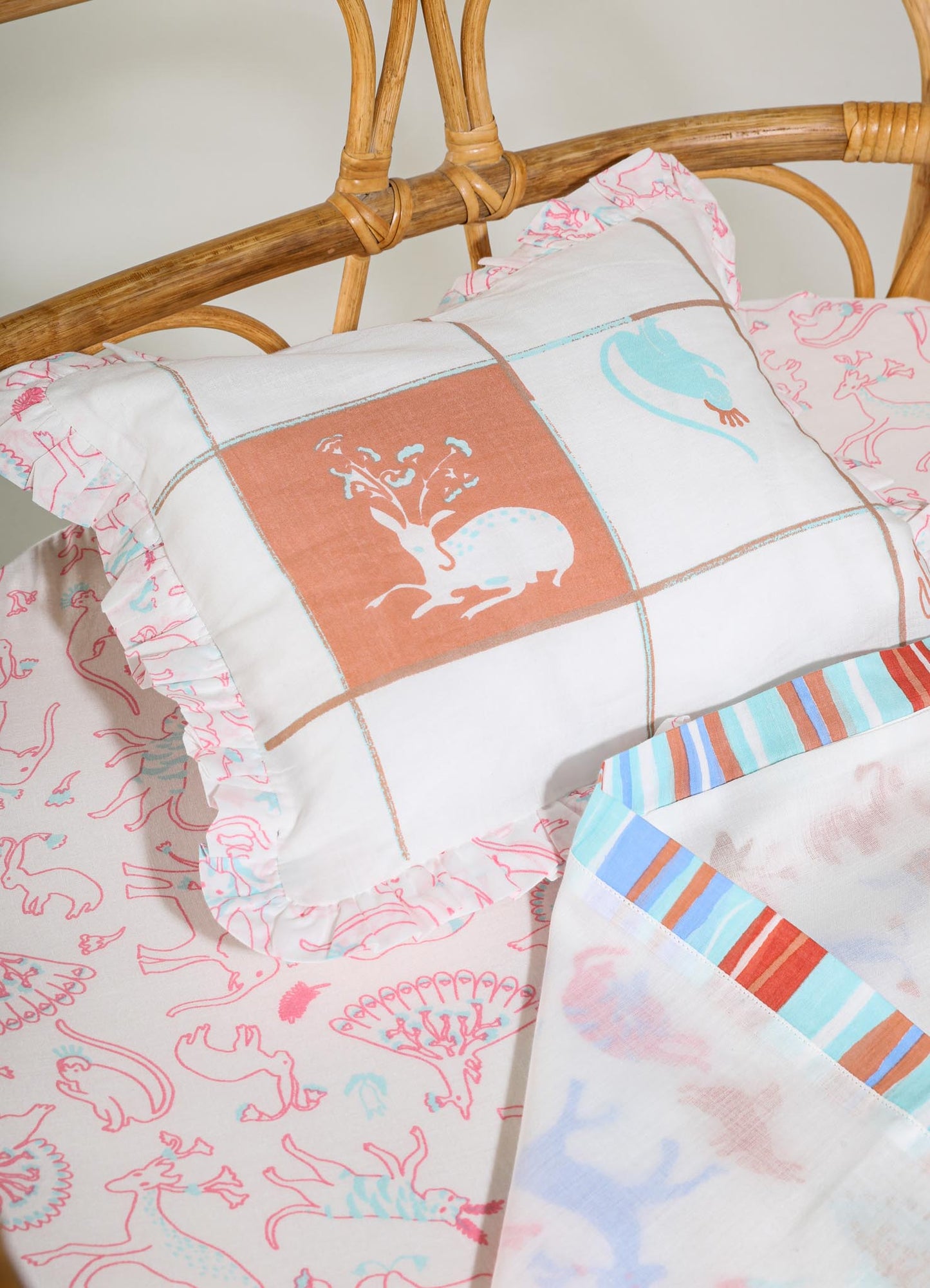Printed cotton bedsheets with whimsical animal and floral motifs in pink and red tones, accompanied by ruffled pillowcases with similar patterning. A rectangular pillow features a stylized embroidered design in pastel blue and brown shades.