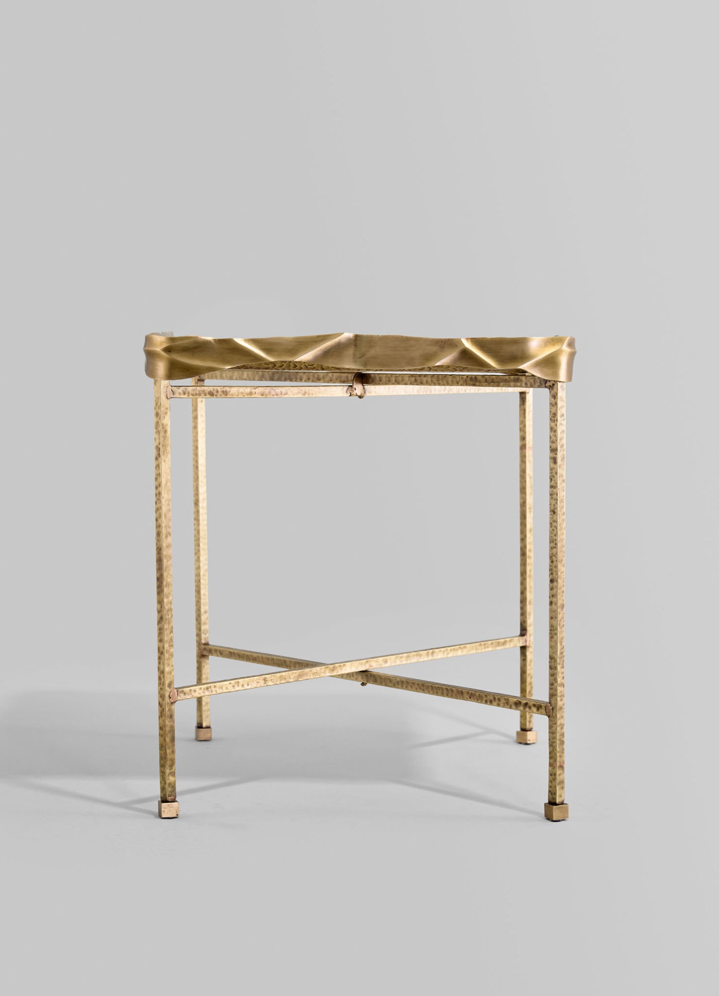 A brass side table with a hammered finish, featuring a sculptural, wavy-edged top surface and a minimalist, geometric frame in a contemporary style.