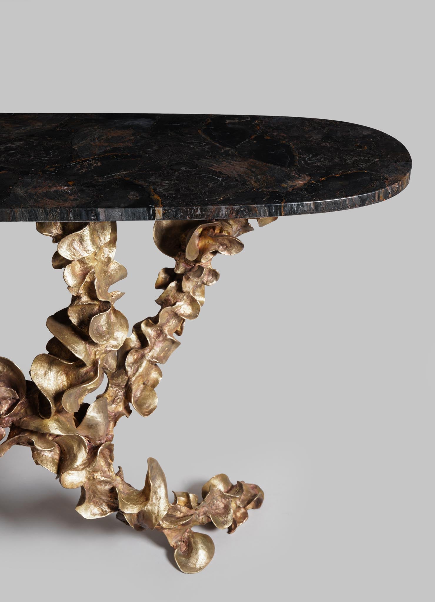 A side table with a round top made of black marble with brown veining, supported by sculptural, gold-colored metal legs with an organic, ruffled appearance.