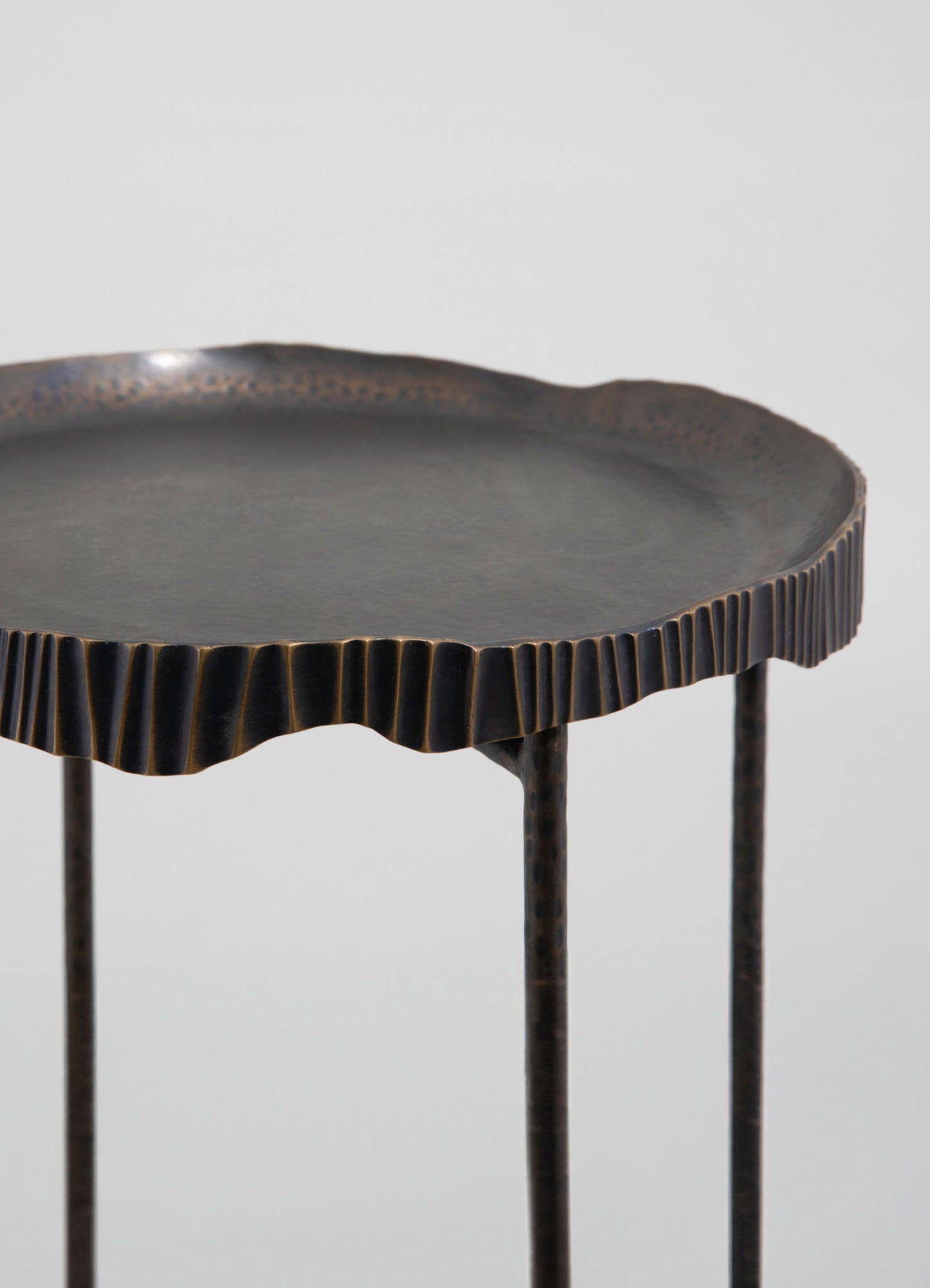 A round metal tray or side table with a textured, ridged edge and a dark, matte finish. The table has slim, straight legs and a minimalist design with a contemporary style.
