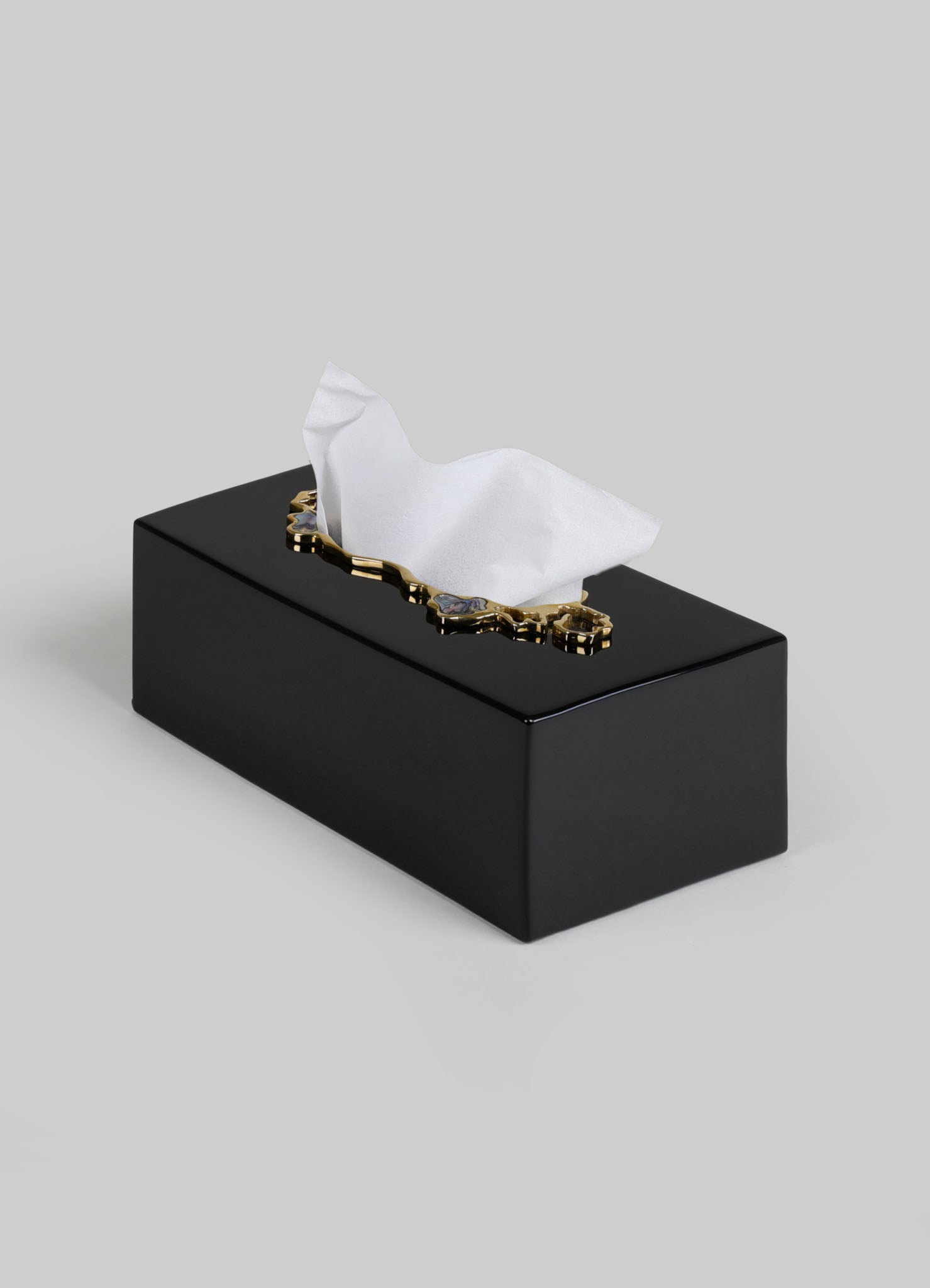 A black rectangular tissue box with a glossy finish, topped with a decorative gold rim and a white tissue partially pulled out.