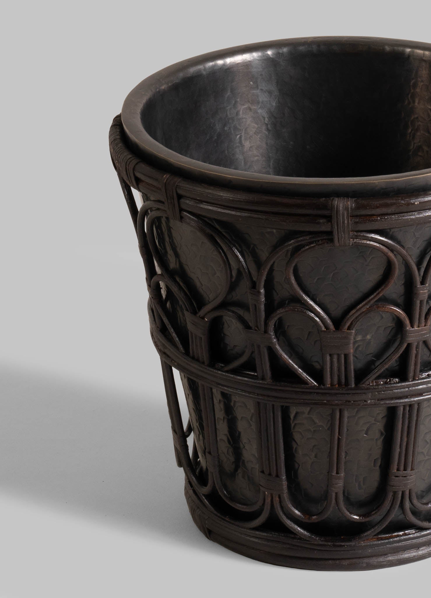 A close-up of the VIYA Manipur Champagne Bucket shows its dark, textured finish and intricate basket-like detailing, inspired by hand-beaten brass. Set against a light gray background, it’s perfect as unique home decor or paired with elegant barware.