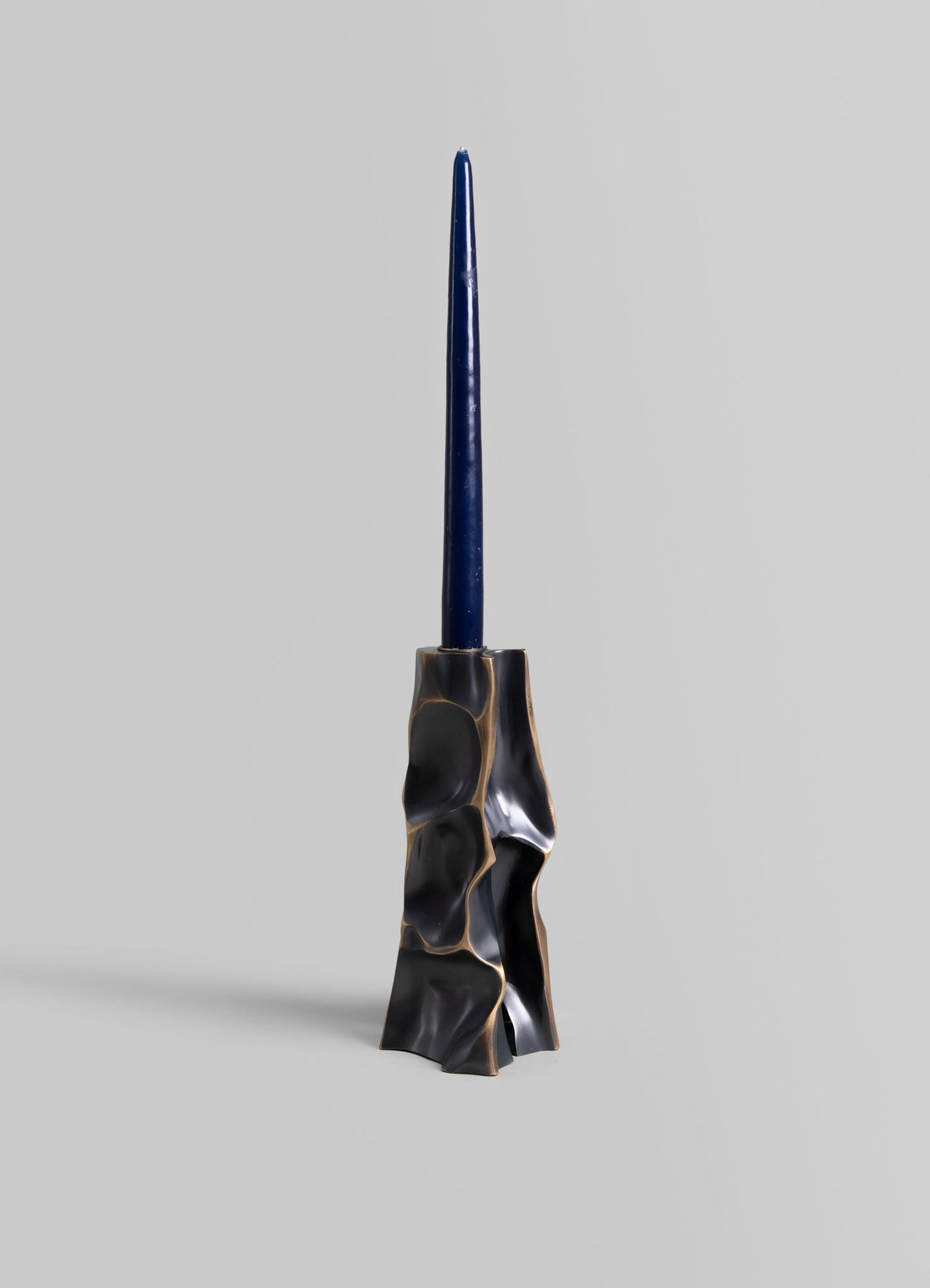 A sculptural candle holder with an irregular, organic-shaped base in black with bronze accents, holding a tall, slender dark blue candle. The design combines contemporary art with decorative functionality.