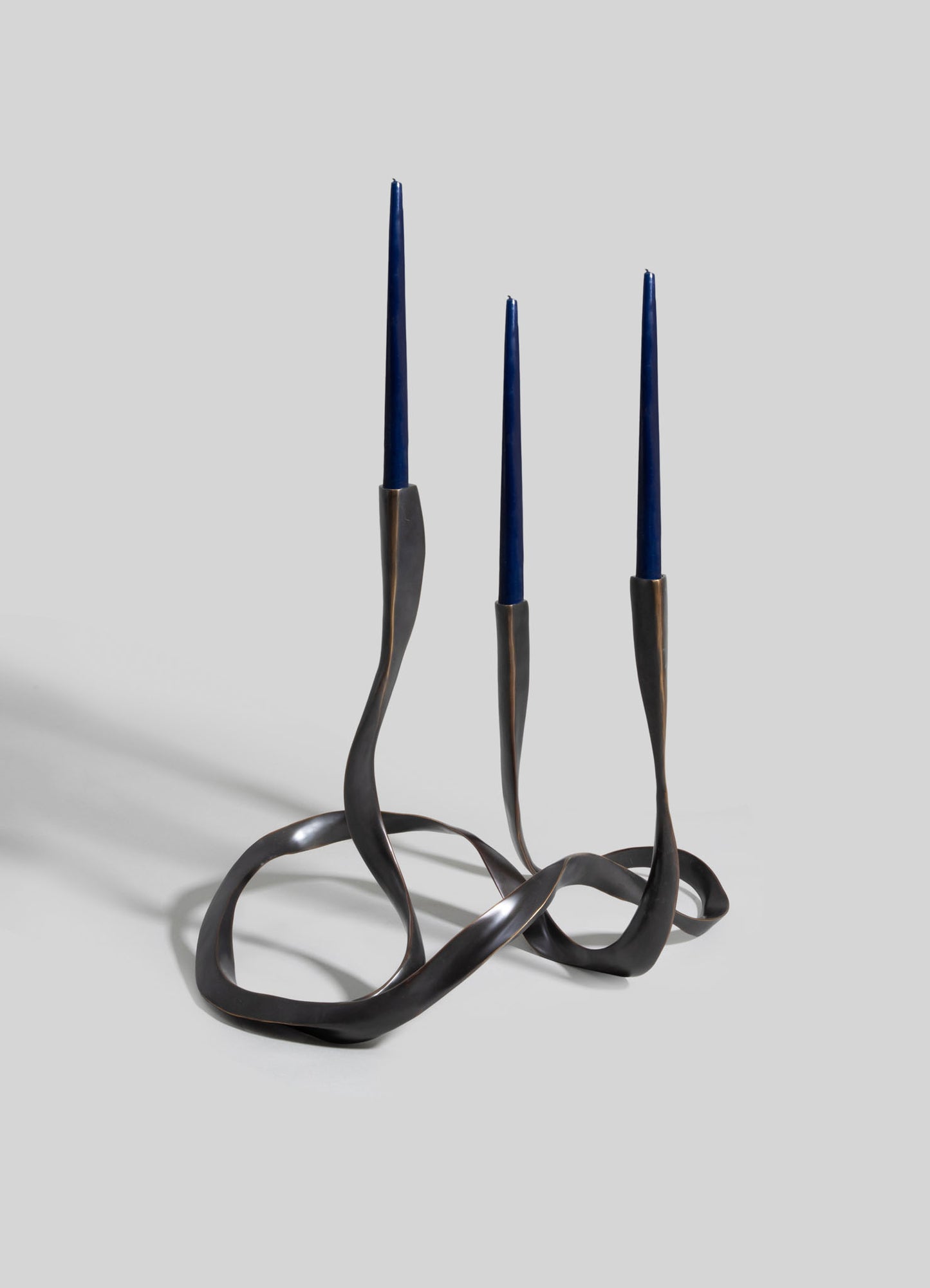 A decorative metal candle holder with three slim navy blue candles, featuring an organic, twisted design in dark bronze or black finish.