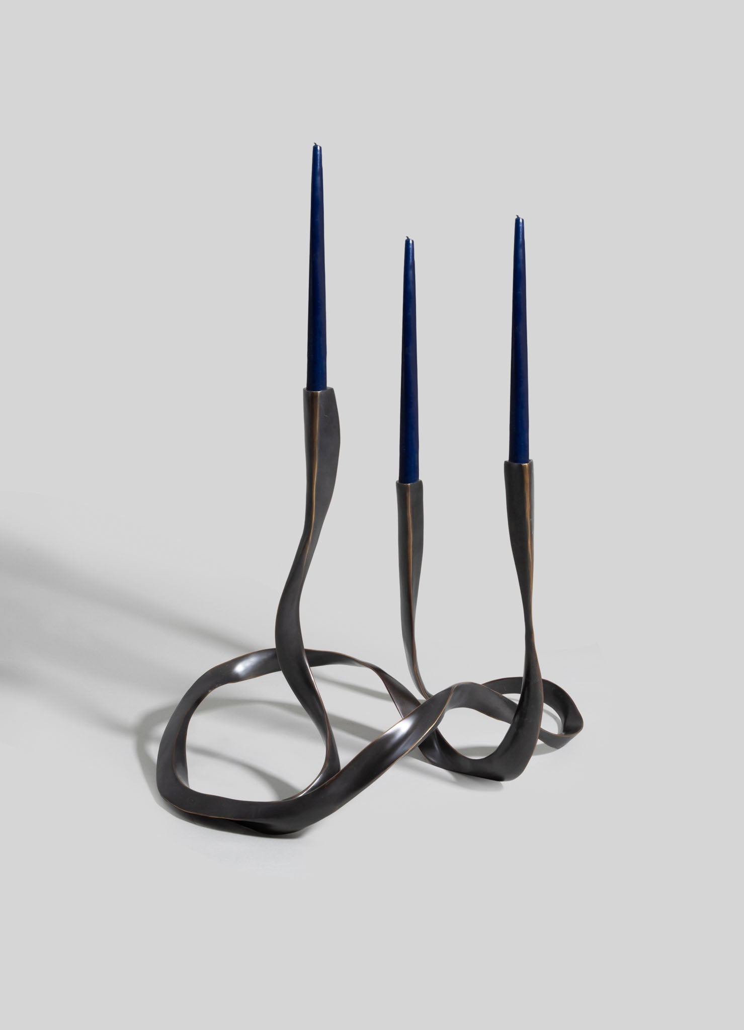A decorative metal candle holder with three slim navy blue candles, featuring an organic, twisted design in dark bronze or black finish.
