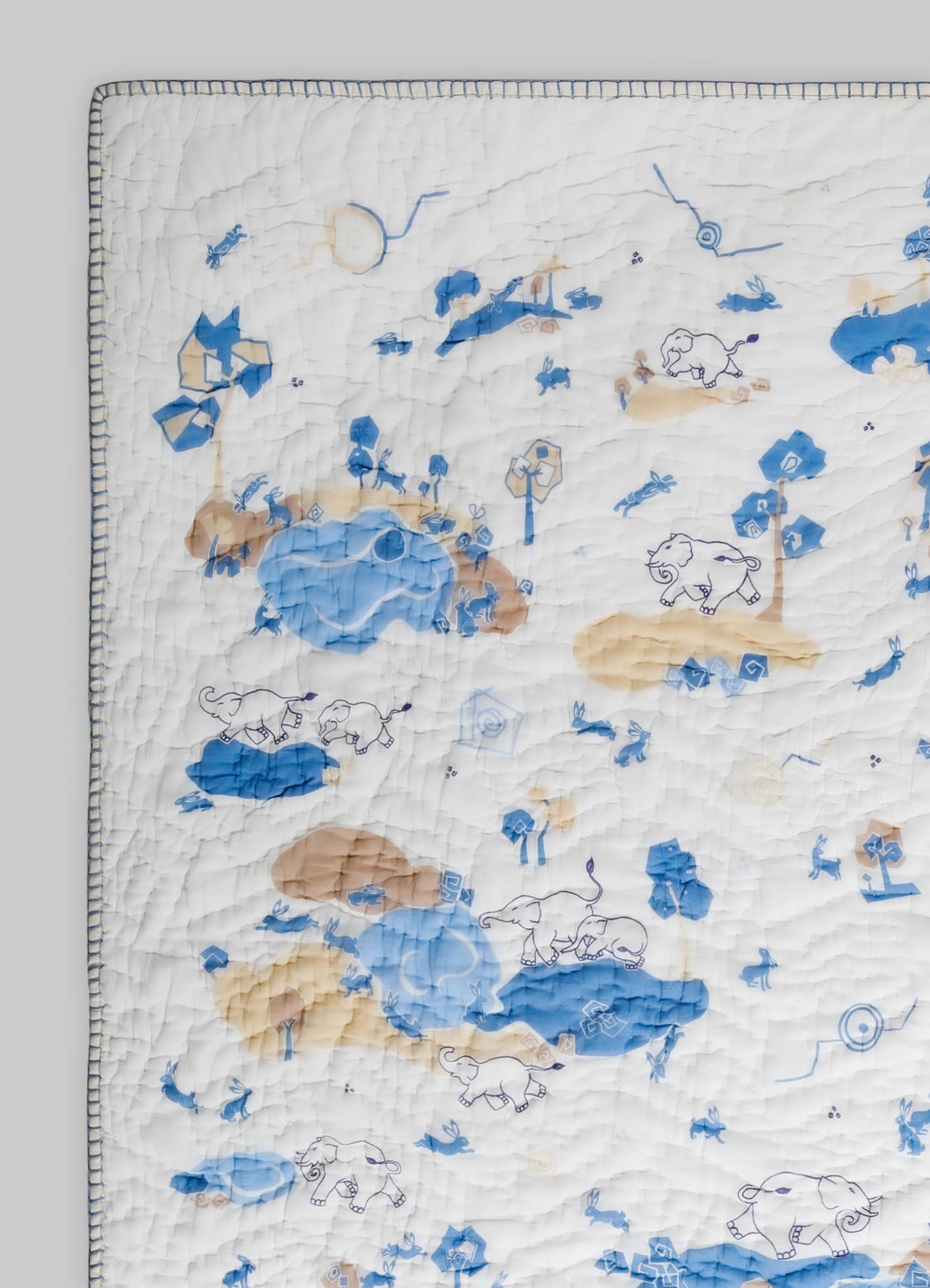 A quilt with a playful animal and nature motif featuring elephants, rabbits, and trees, in shades of blue, beige, and white. The quilt has a stitched texture with a decorative border around the edges.