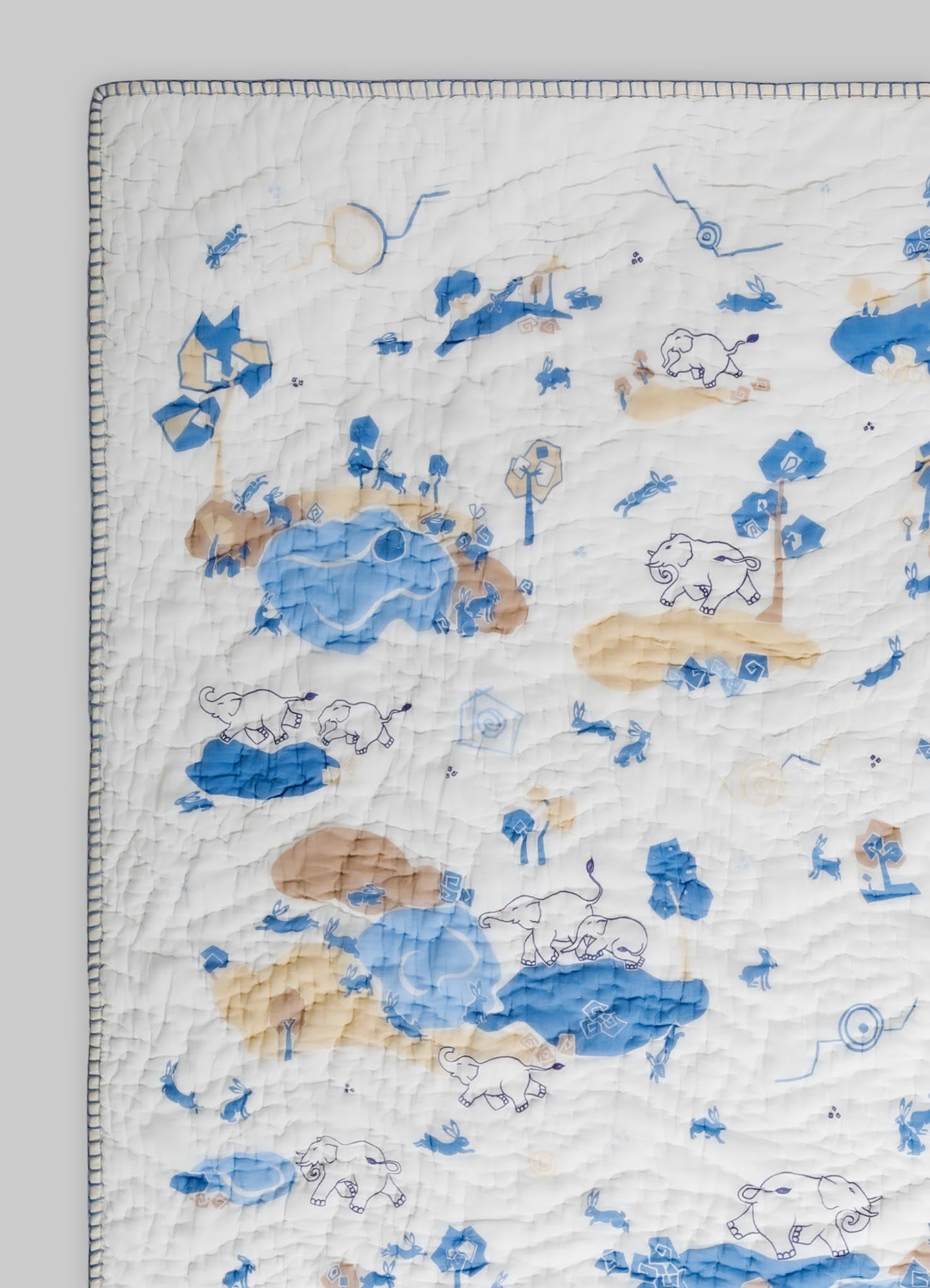 A quilt with a playful animal and nature motif featuring elephants, rabbits, and trees, in shades of blue, beige, and white. The quilt has a stitched texture with a decorative border around the edges.