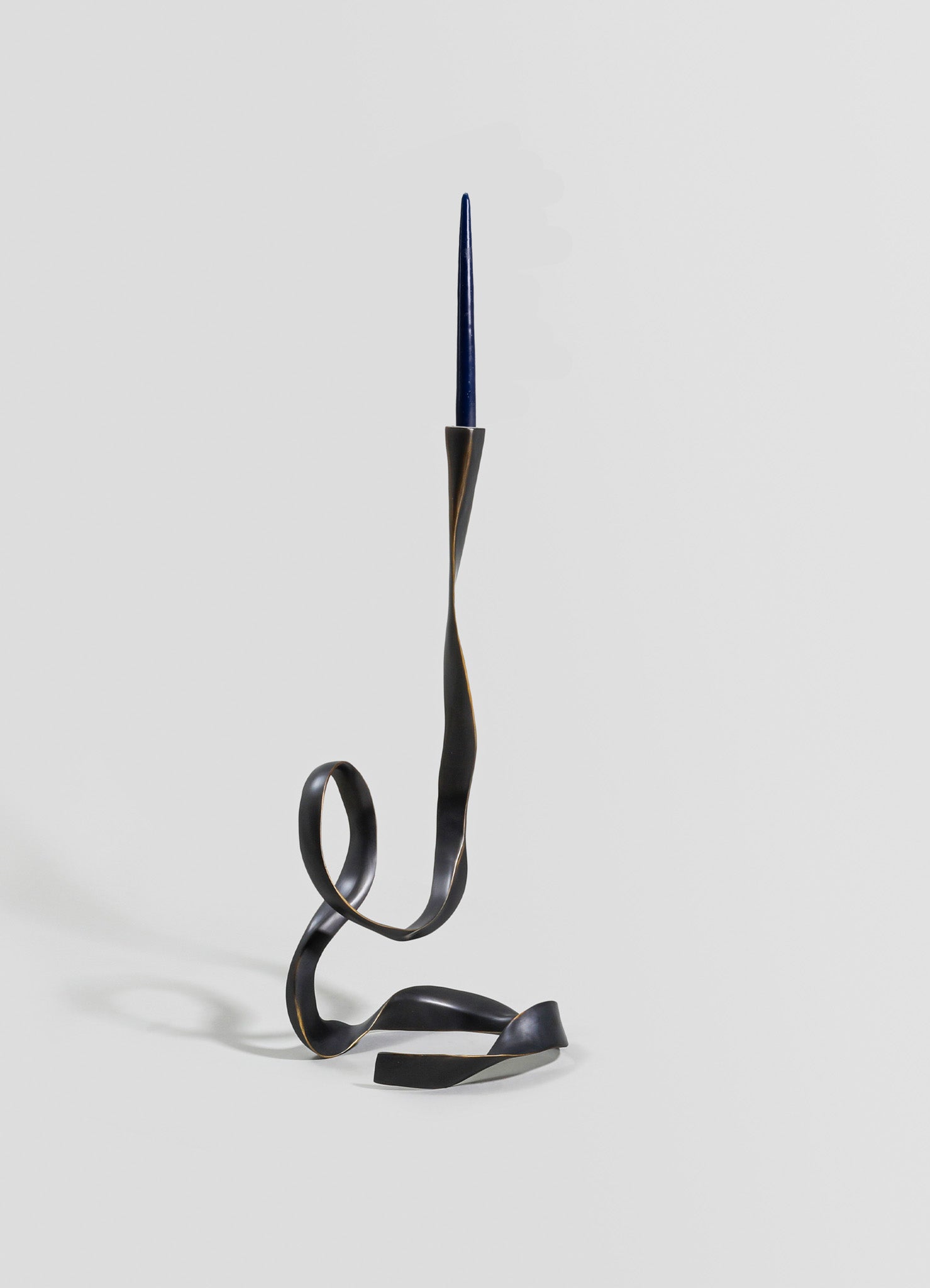 A sculptural candle holder with a black metal finish, featuring a twisted, ribbon-like design and a single blue taper candle inserted into the top. The piece has a contemporary, artistic style with an abstract, fluid form.