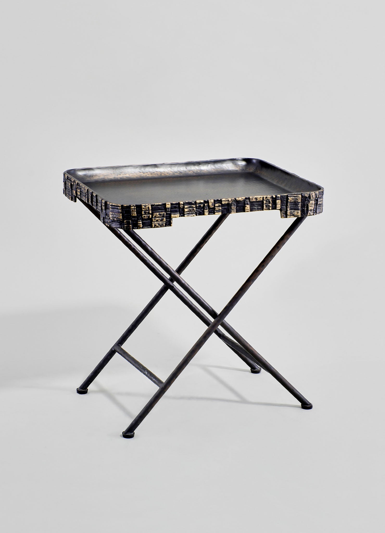 A metal folding serving tray table with a rectangular black tray featuring a textured, geometric patterned border in dark tones, supported by crossed metal legs.