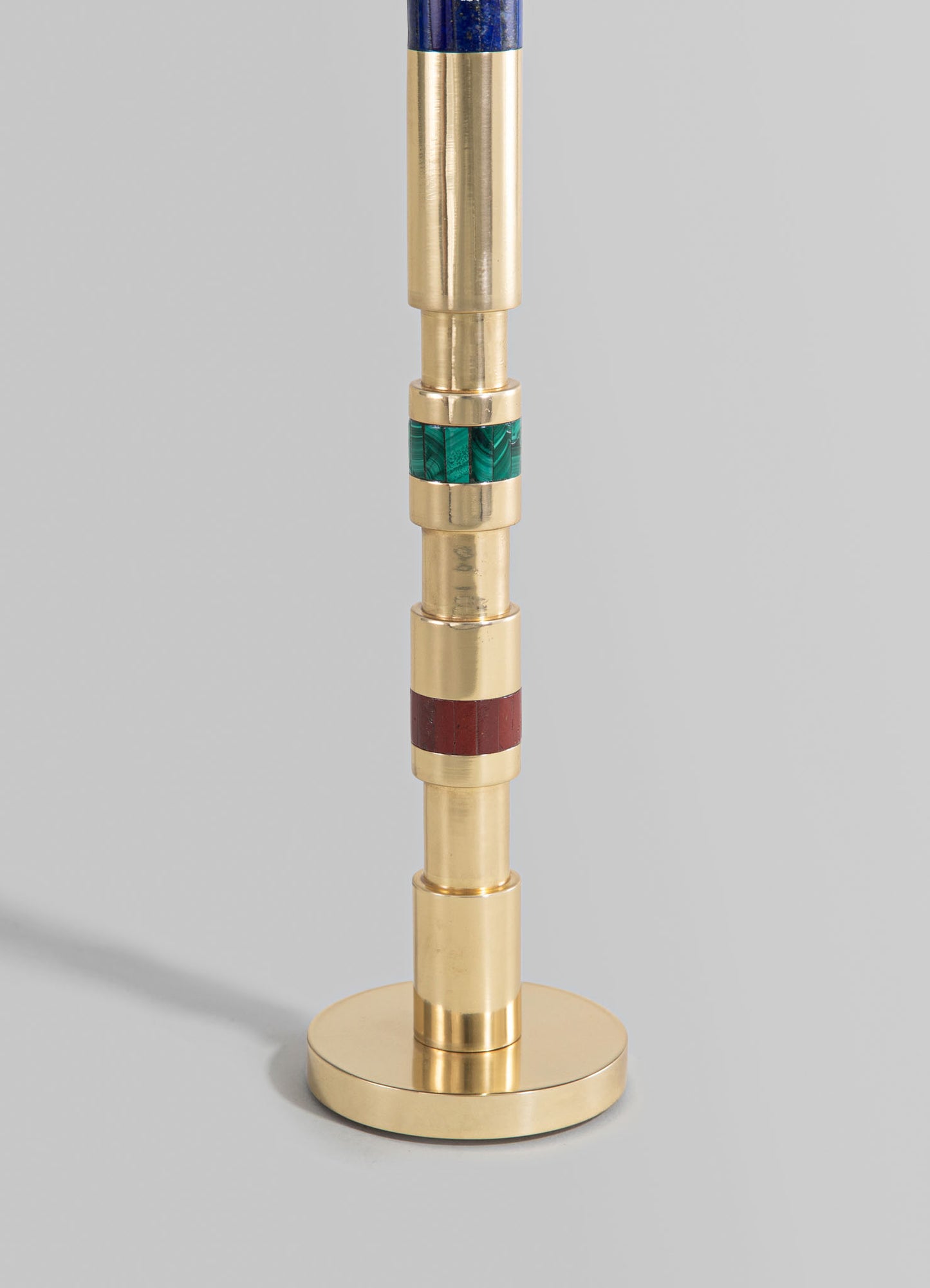 A brass candlestick with a cylindrical shape, featuring colored inlay bands in green, red, and blue, mounted on a round base with a modern, minimalist design.