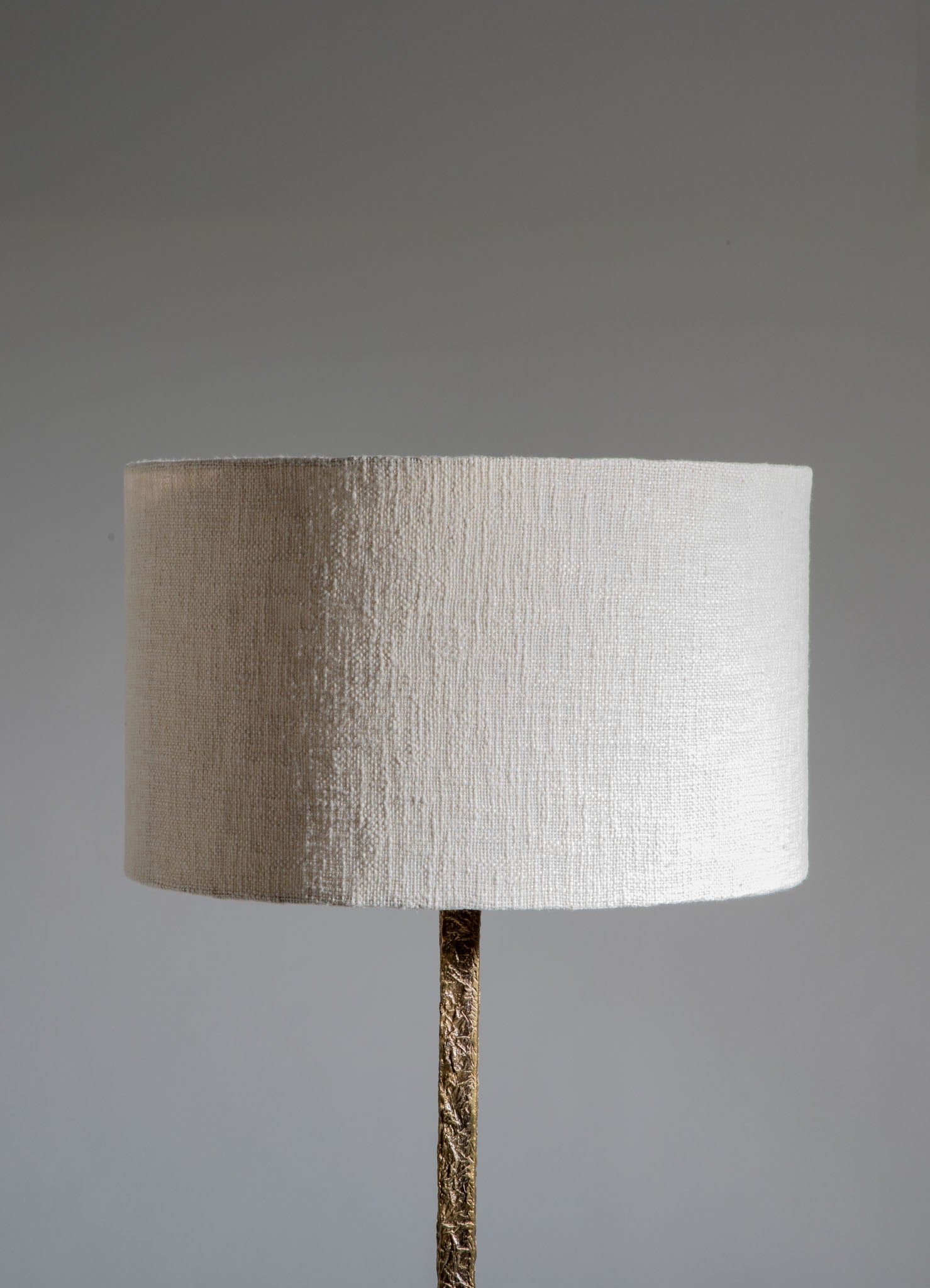 A table lamp featuring a textured fabric cylindrical shade in a neutral beige tone, mounted on a metallic gold base with an intricate, embossed pattern.