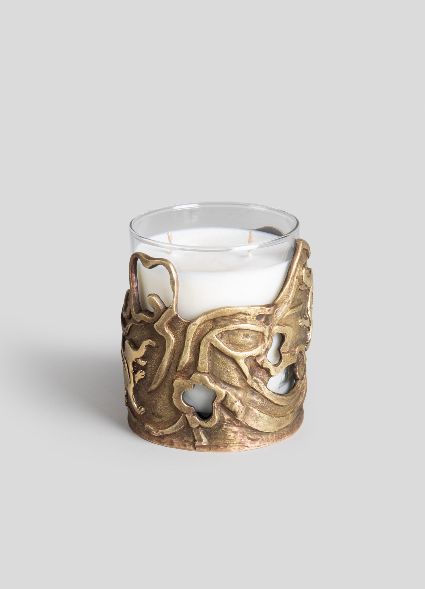 A glass candle holder wrapped in an ornate, crafted metal sleeve with an abstract organic motif in antique gold finish, holding a white pillar candle.