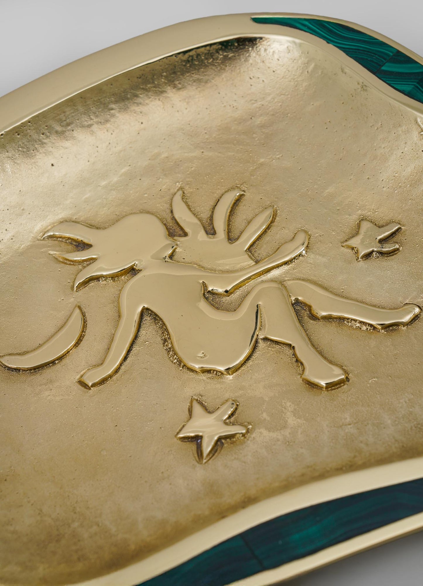 A decorative tray with a gold finish featuring a raised fairy motif and small stars, accented with a strip of green malachite or similar material along the edge. The tray's surface has a textured, matte background contrasting with the shiny, embossed design.