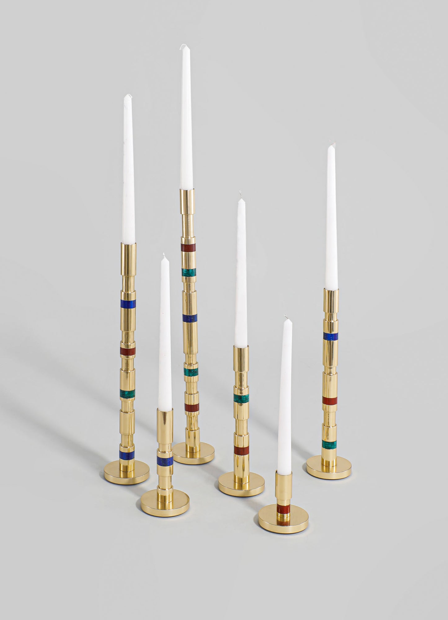 Set of six taper candles with white wax and metal holders featuring colorful, geometric bands in red, blue, and green, with a gold-tone finish. The candles are tall and slender, arranged on a plain background.