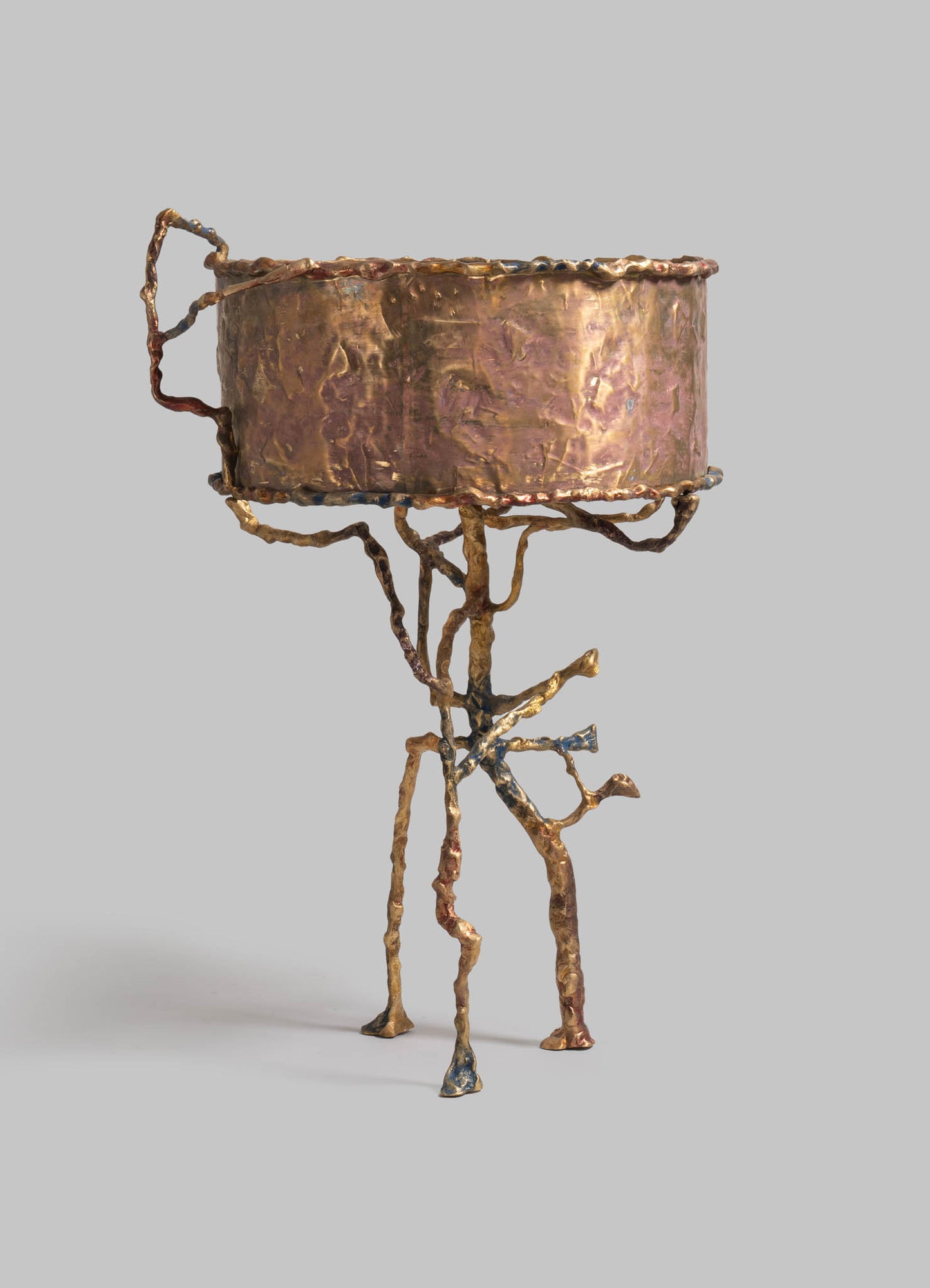 A sculpture with a metallic finish resembling bronze or copper, featuring a cylindrical top with an uneven, textured surface, supported by an abstract, branching base with a rough, organic appearance.