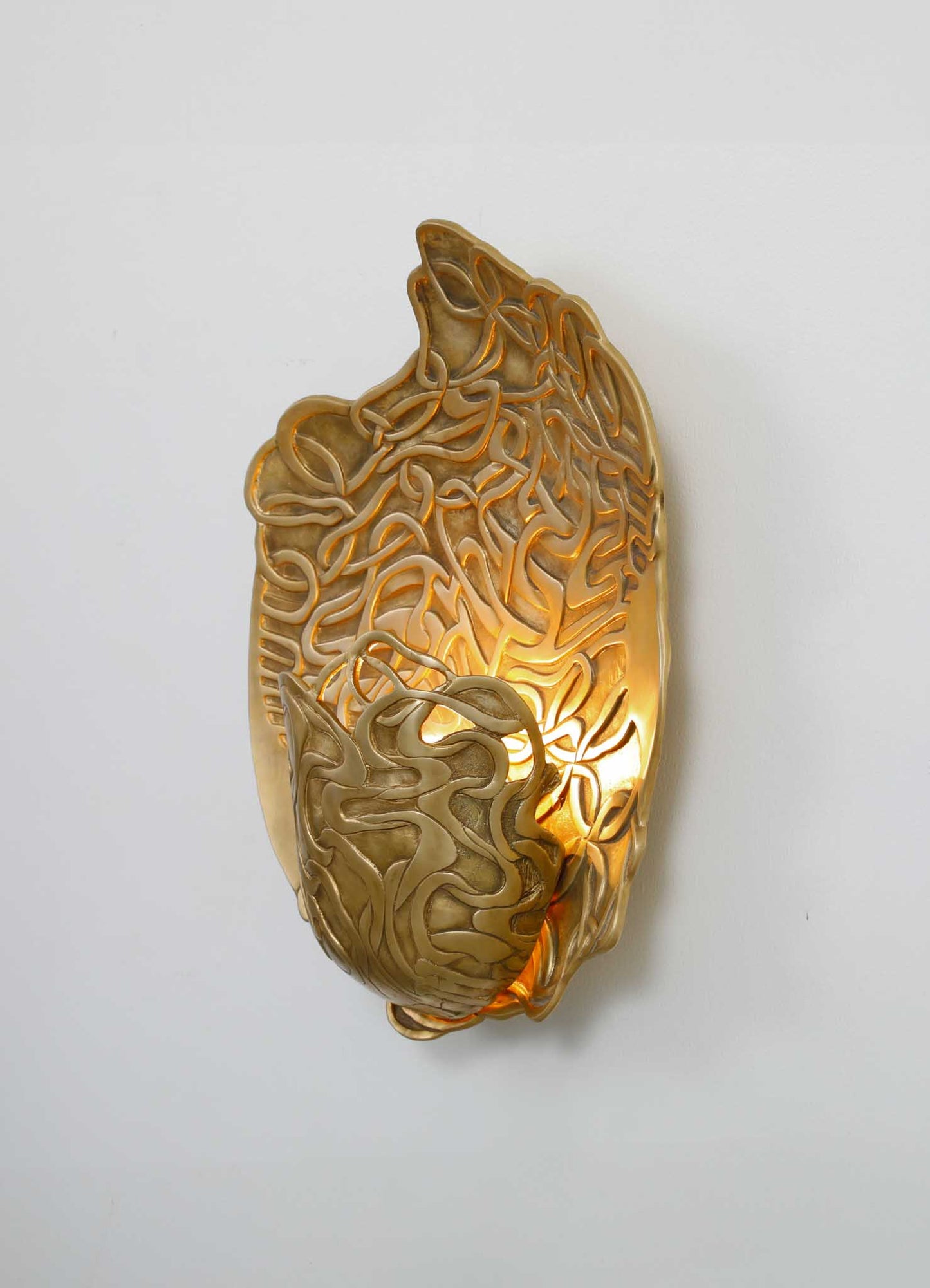 A wall-mounted sconce made of gold-colored metal with an intricate, organic, swirling motif. The design resembles a stylized leaf or floral pattern, creating a decorative and luminous accent in a contemporary or luxury interior setting.