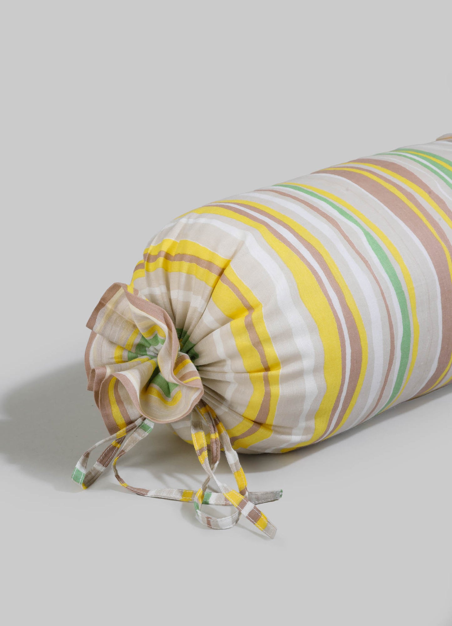 Striped fabric bolster pillow with pastel yellow, pink, green, and beige tones, featuring a ruffled end and tied bows on a light-colored surface.
