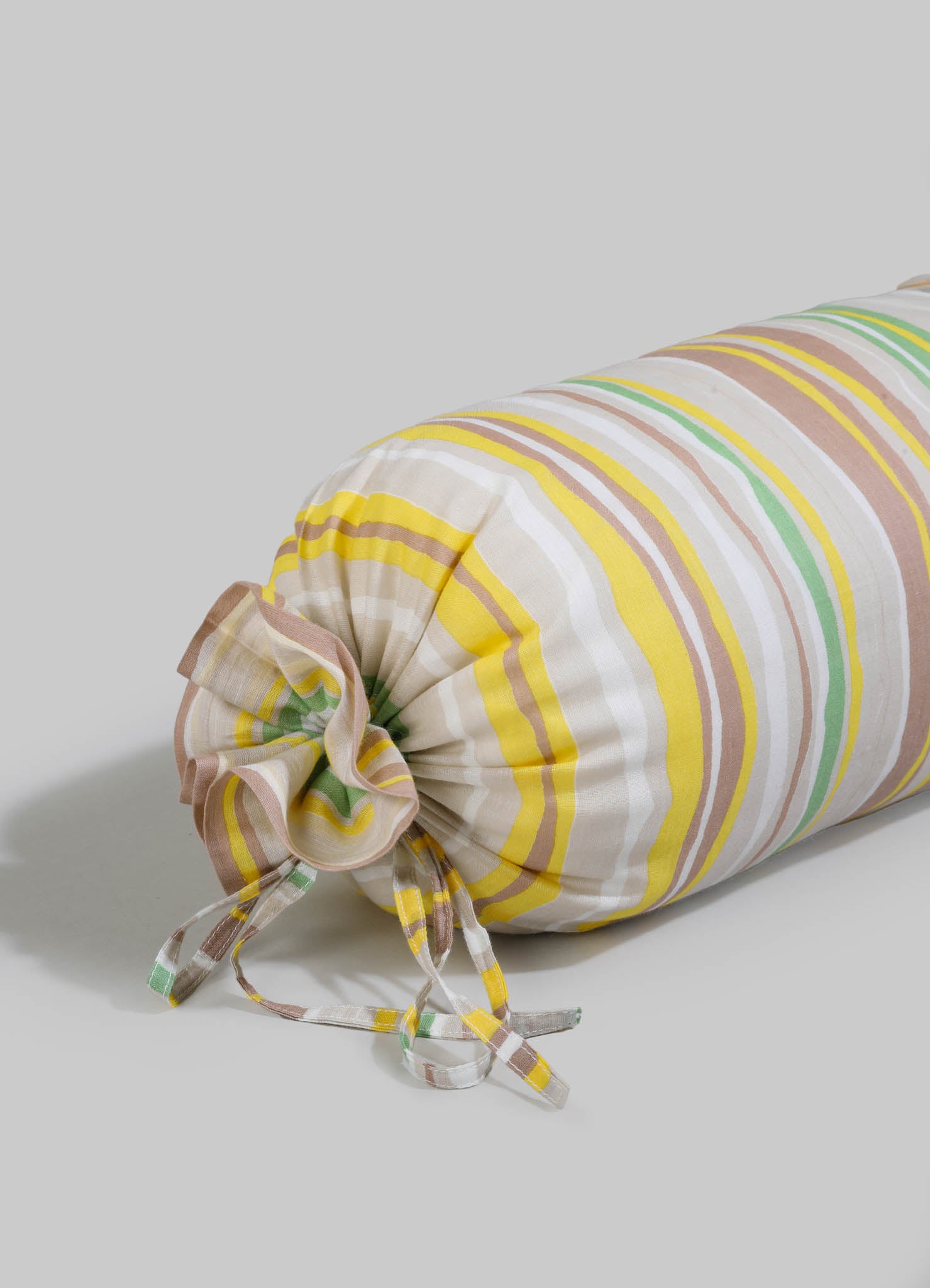 Striped fabric bolster pillow with pastel yellow, pink, green, and beige tones, featuring a ruffled end and tied bows on a light-colored surface.