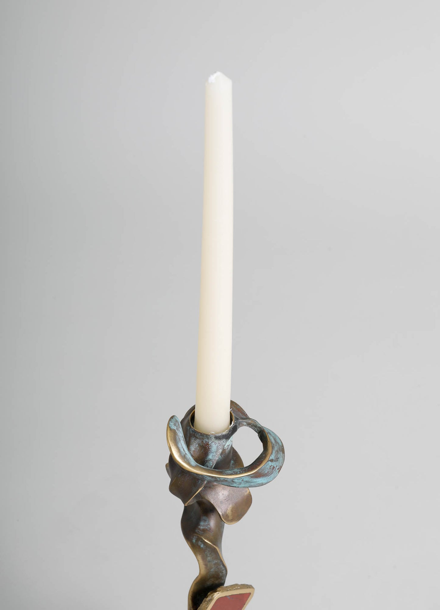A metallic candle holder with an abstract, sculptural design in aged brass tones, holding a tall, white taper candle. The piece features organic, flowing curves with a textured surface, presented against a plain gray background.