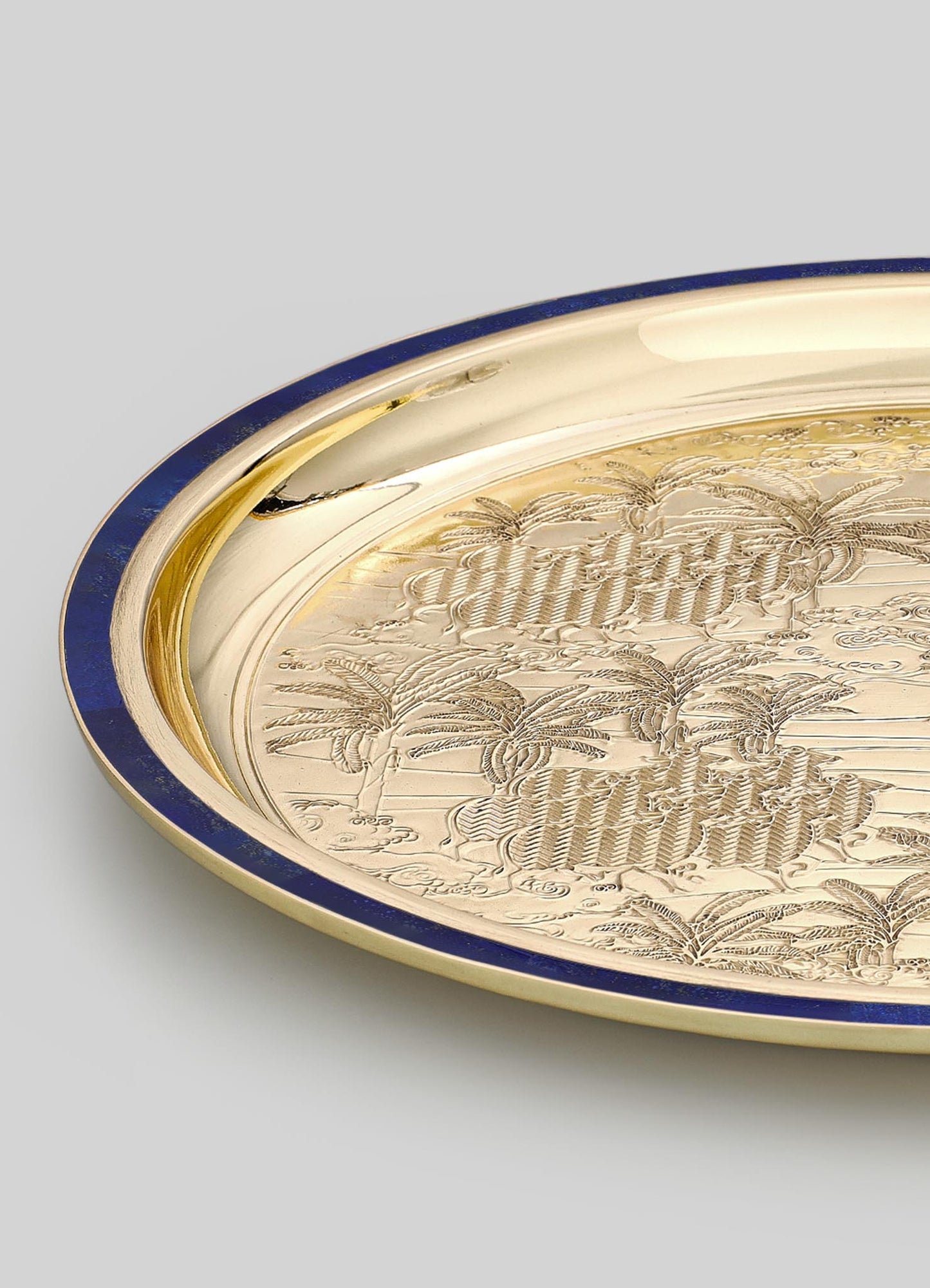 A round decorative tray with a polished gold-tone metal surface, featuring engraved palm tree motifs and the word "Hawaii," bordered with a dark blue rim.