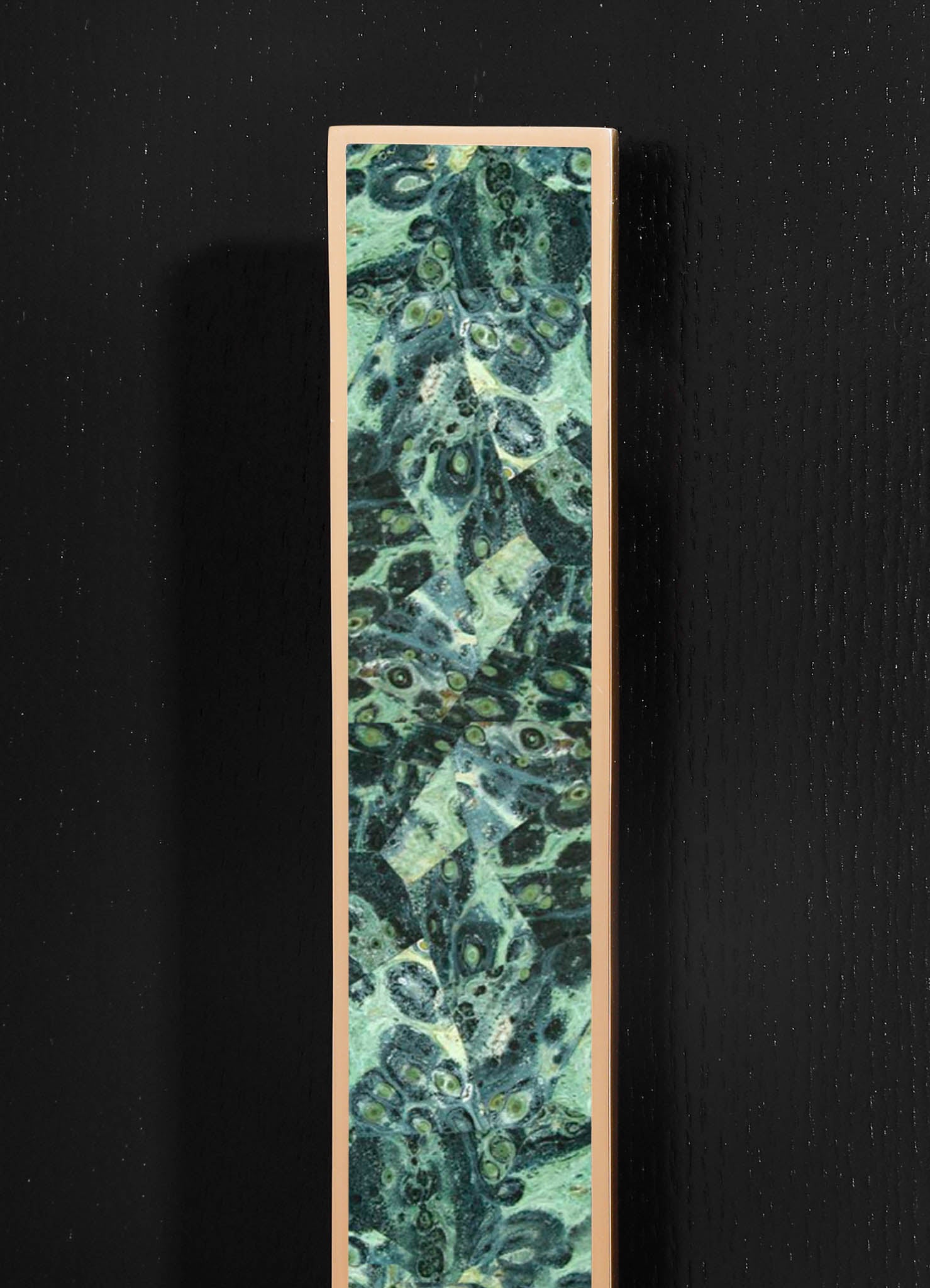 A tall, rectangular mirror with a gold or brass frame featuring a vibrant, abstract pattern in shades of green, teal, and black, mounted on a black textured wall.