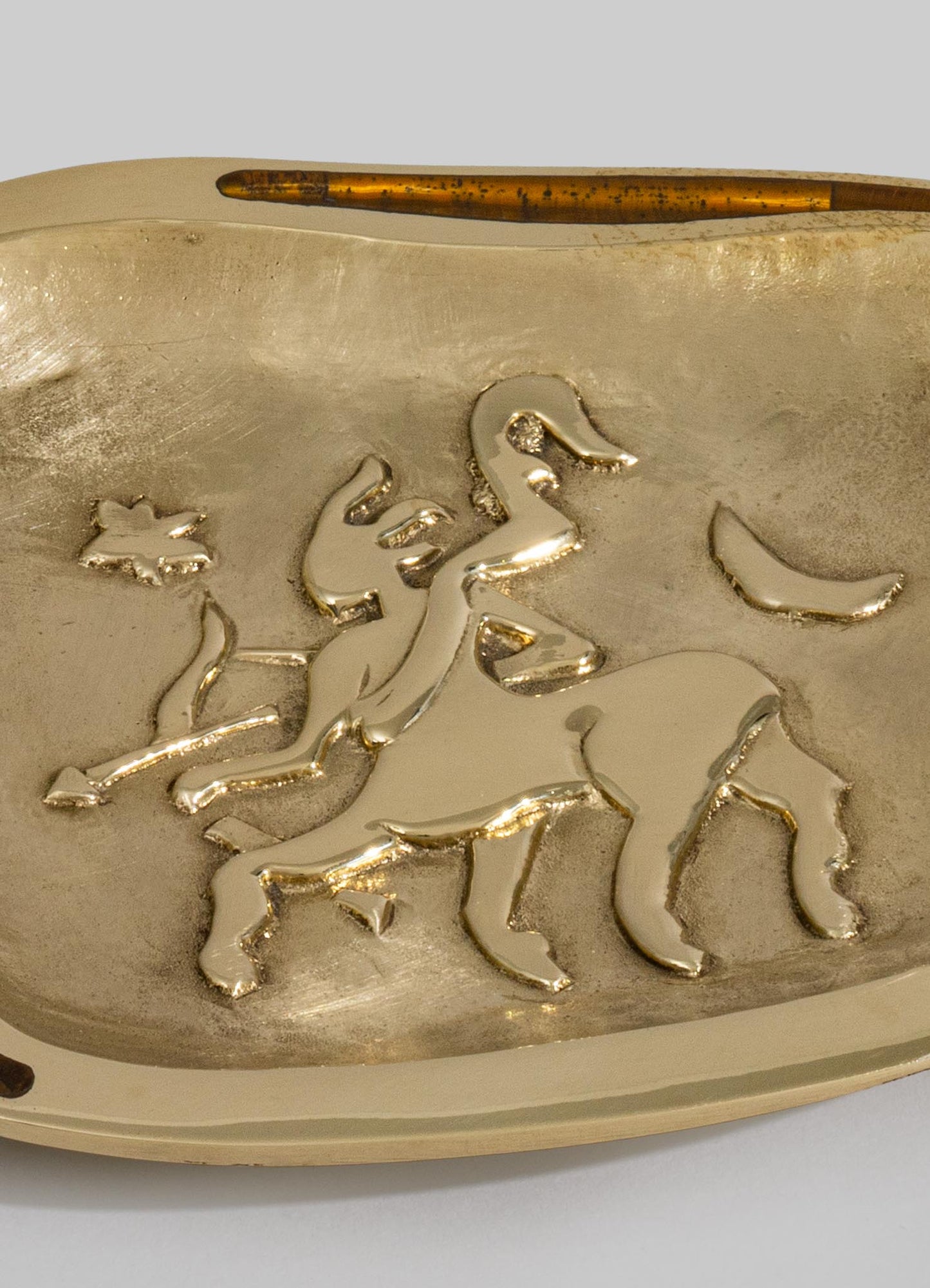 A decorative brass tray with an embossed horse motif, featuring a polished gold finish and organic, flowing lines. The tray has a rounded edge and a textured surface, conveying an artisanal, handcrafted style.