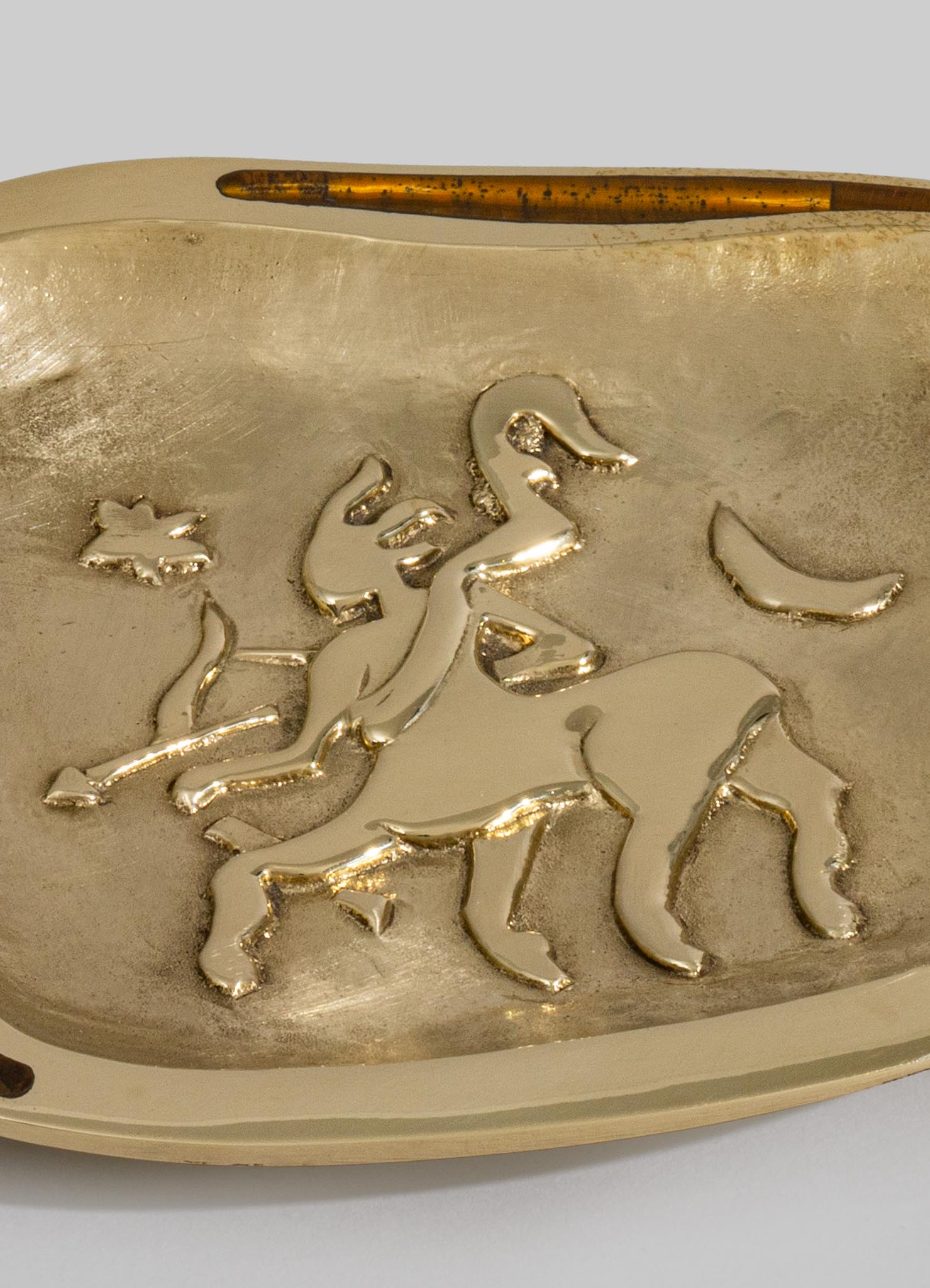 A decorative brass tray with an embossed horse motif, featuring a polished gold finish and organic, flowing lines. The tray has a rounded edge and a textured surface, conveying an artisanal, handcrafted style.