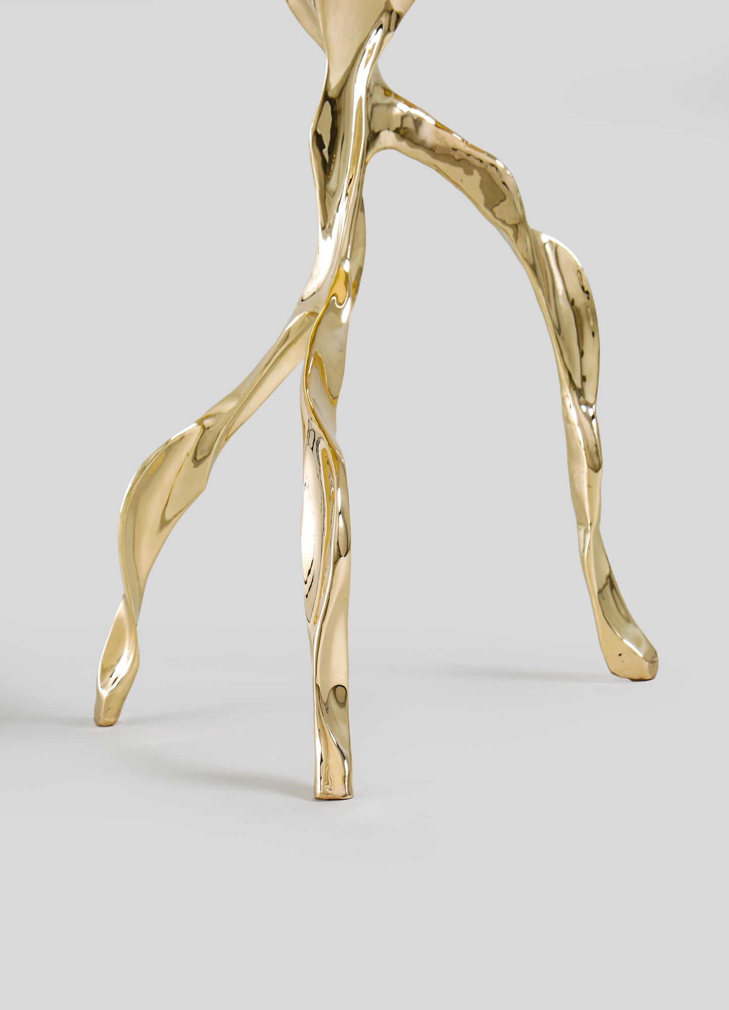A close-up of VIYA’s Under The Banyan Gold Side Table Large, a sculptural, three-legged metallic piece with glossy, organic forms reminiscent of aerial roots, set against a plain light gray background.
