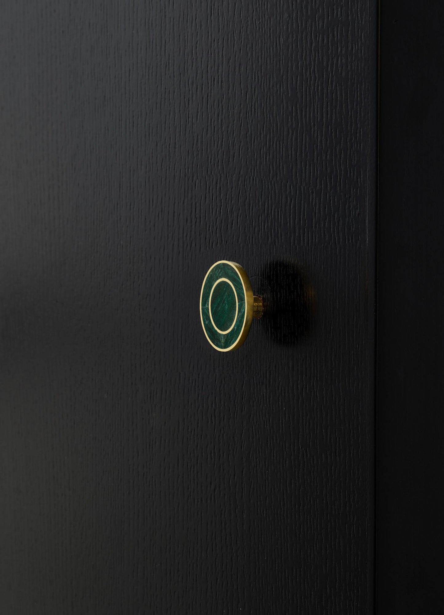 Close-up of a black textured wooden surface with a small, round brass and green marble drawer knob displaying a circular pattern. The style appears modern and minimalist.