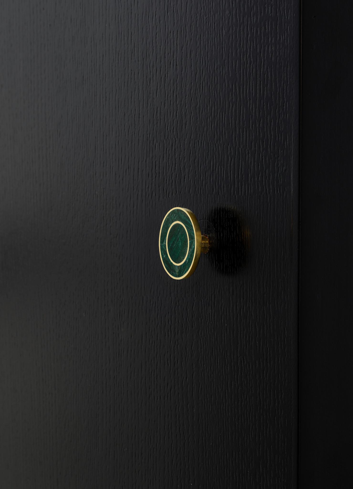 Close-up of a black textured wooden surface with a small, round brass and green marble drawer knob displaying a circular pattern. The style appears modern and minimalist.