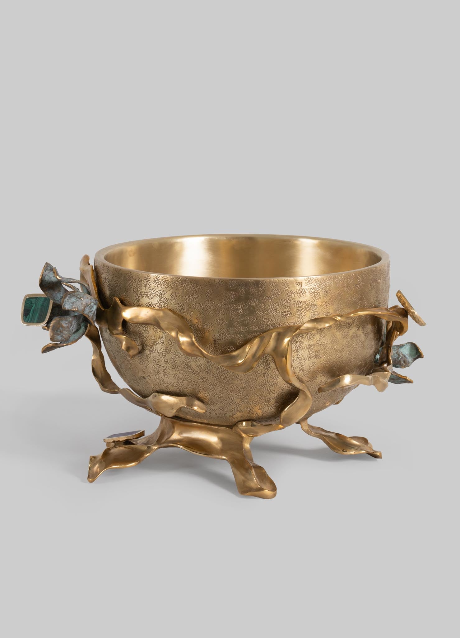 A decorative gold bowl with a textured surface, featuring an organic design with twisted metallic branches and floral motifs in greenish and bluish tones, mounted on a sculptural base.